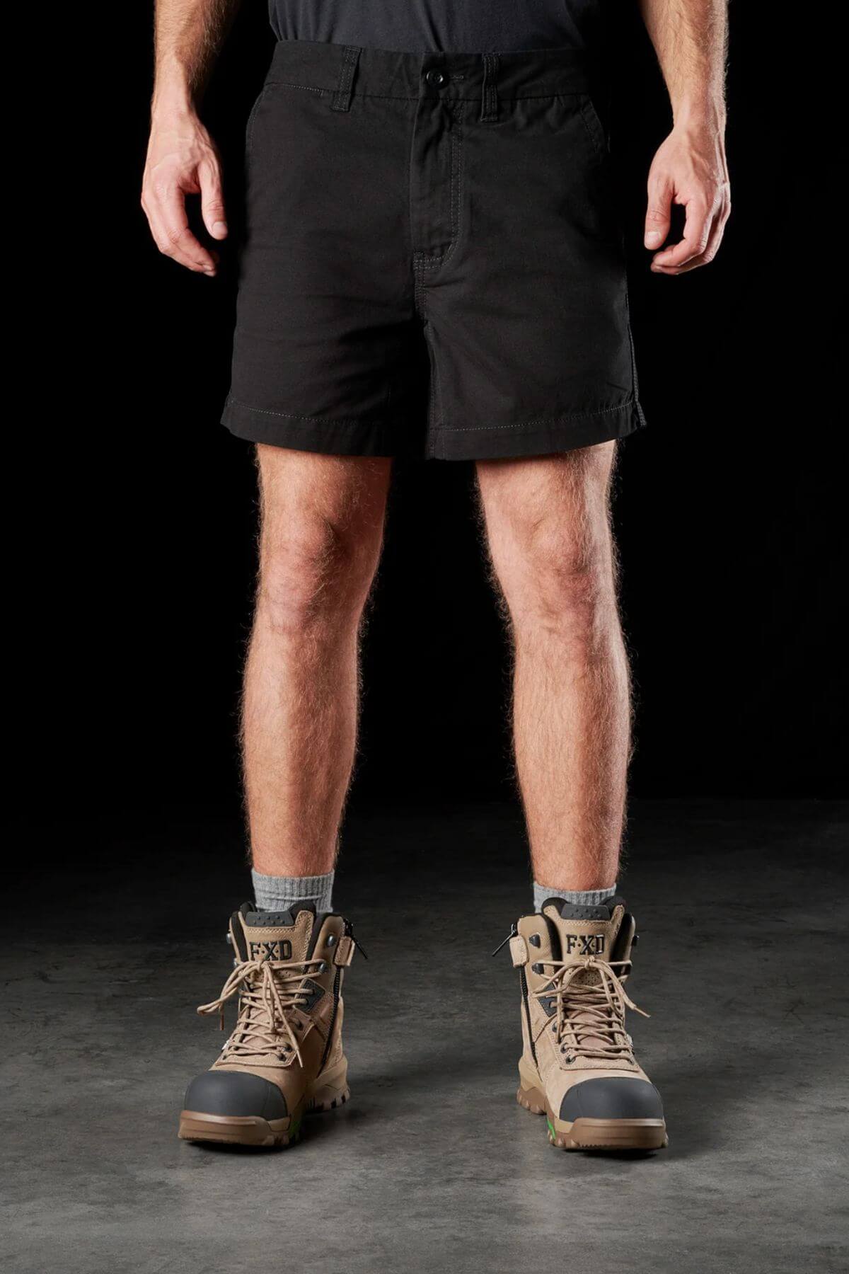 A person is standing with their legs visible, wearing black FXD WS-2 Short Shorts made from cotton twill. They have gray socks and sturdy brown work boots labeled "FXD." The person is also wearing a black shirt, though only part of it is visible. The background is dark.