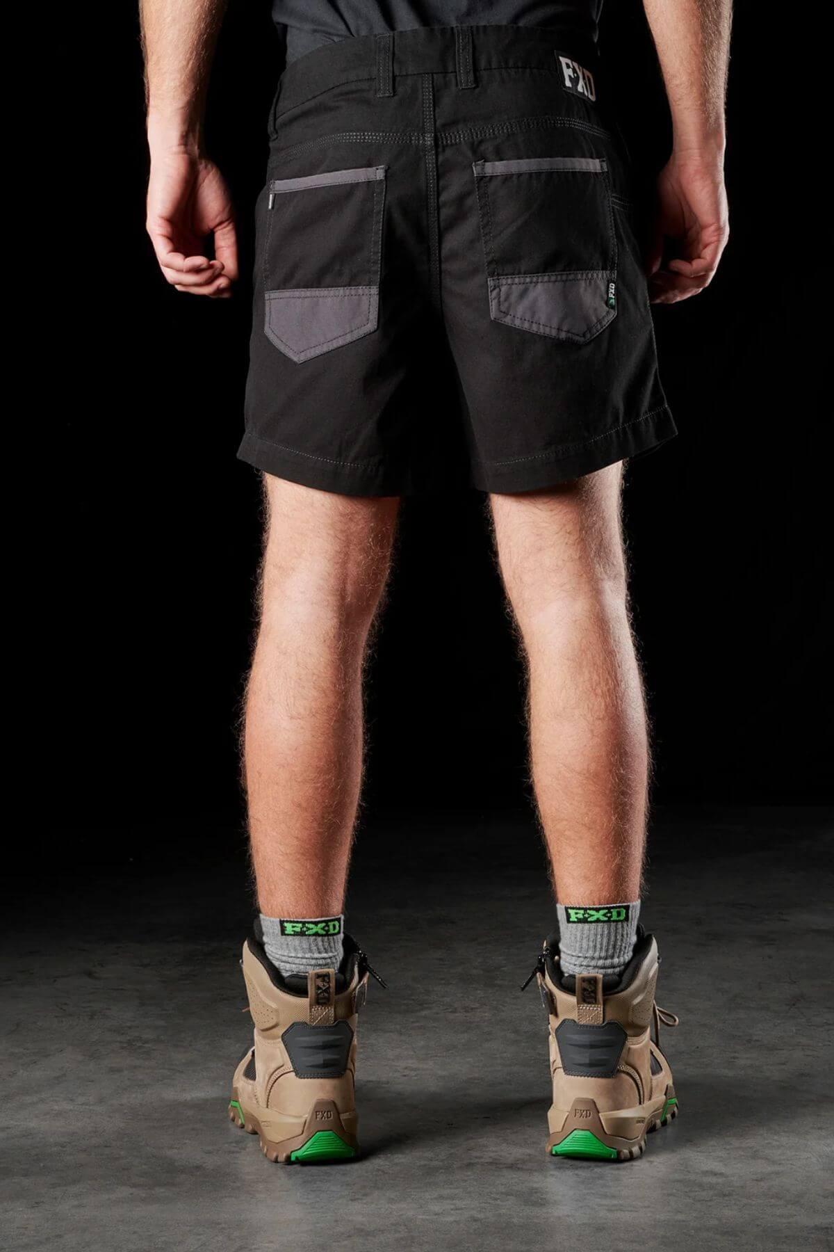 A person stands with their back to the camera, wearing black FXD WS-2 Short Shorts featuring gray pockets and a belt. They are also sporting brown hiking boots with green accents while standing on a gray surface against a black background.
