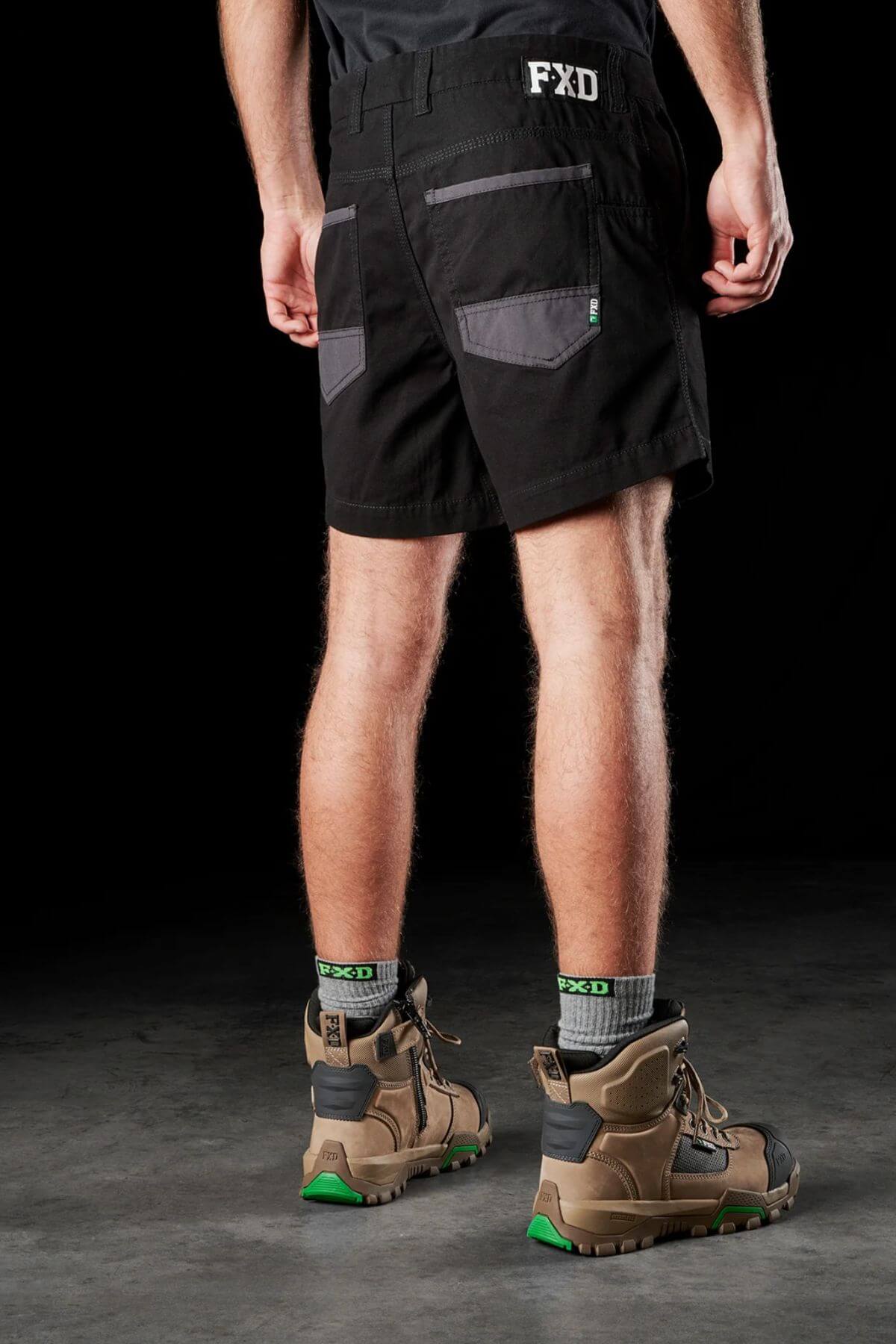 A person is standing with their back to the camera, wearing FXD WS-2 Short Shorts made of black cotton twill with gray pockets. They are also sporting beige hiking boots with green accents and gray socks that say "FXD." The dark background highlights the lower half of their body.