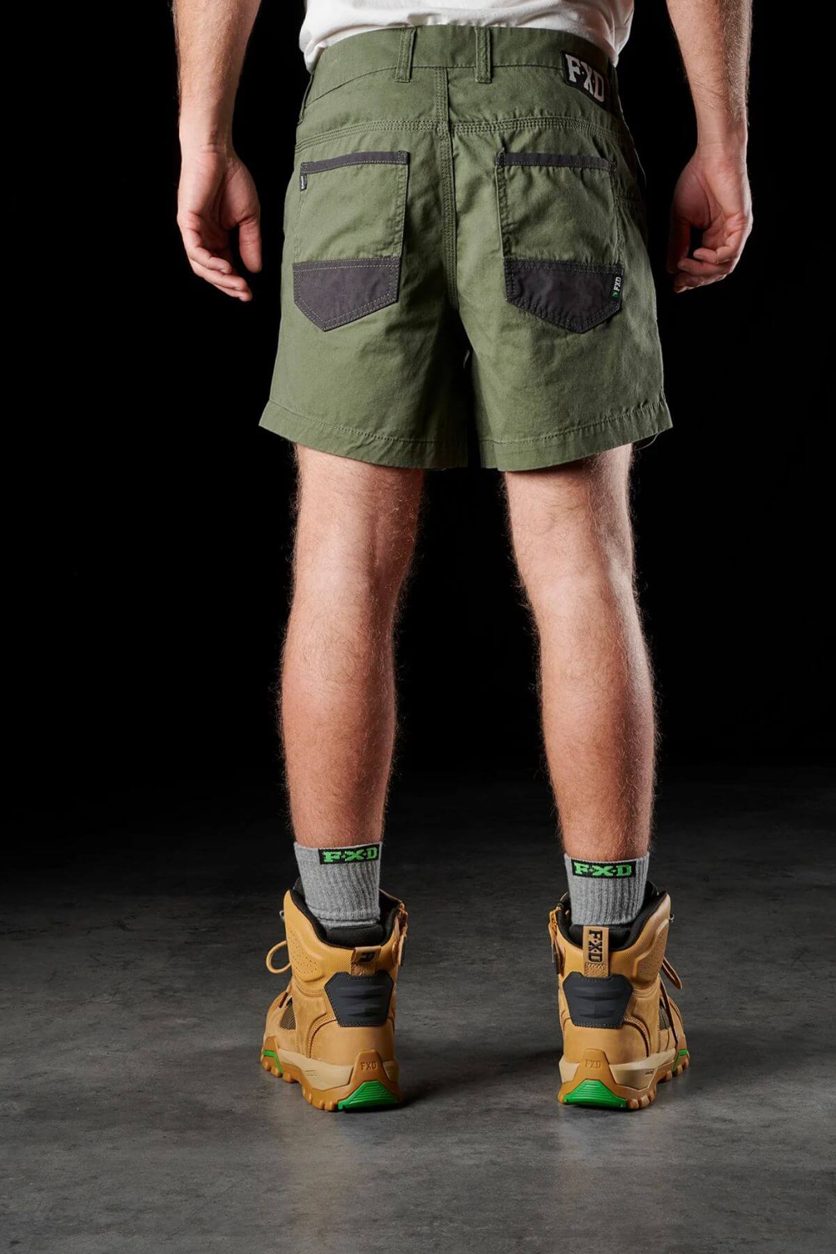Rear view of a person wearing green FXD WS-2 Short Shorts, which have two pockets and an FXD logo, paired with gray socks and tan work boots featuring green soles. The shorts are made from soft touch cotton twill and include DURA500™ abrasion panels. The background is black.