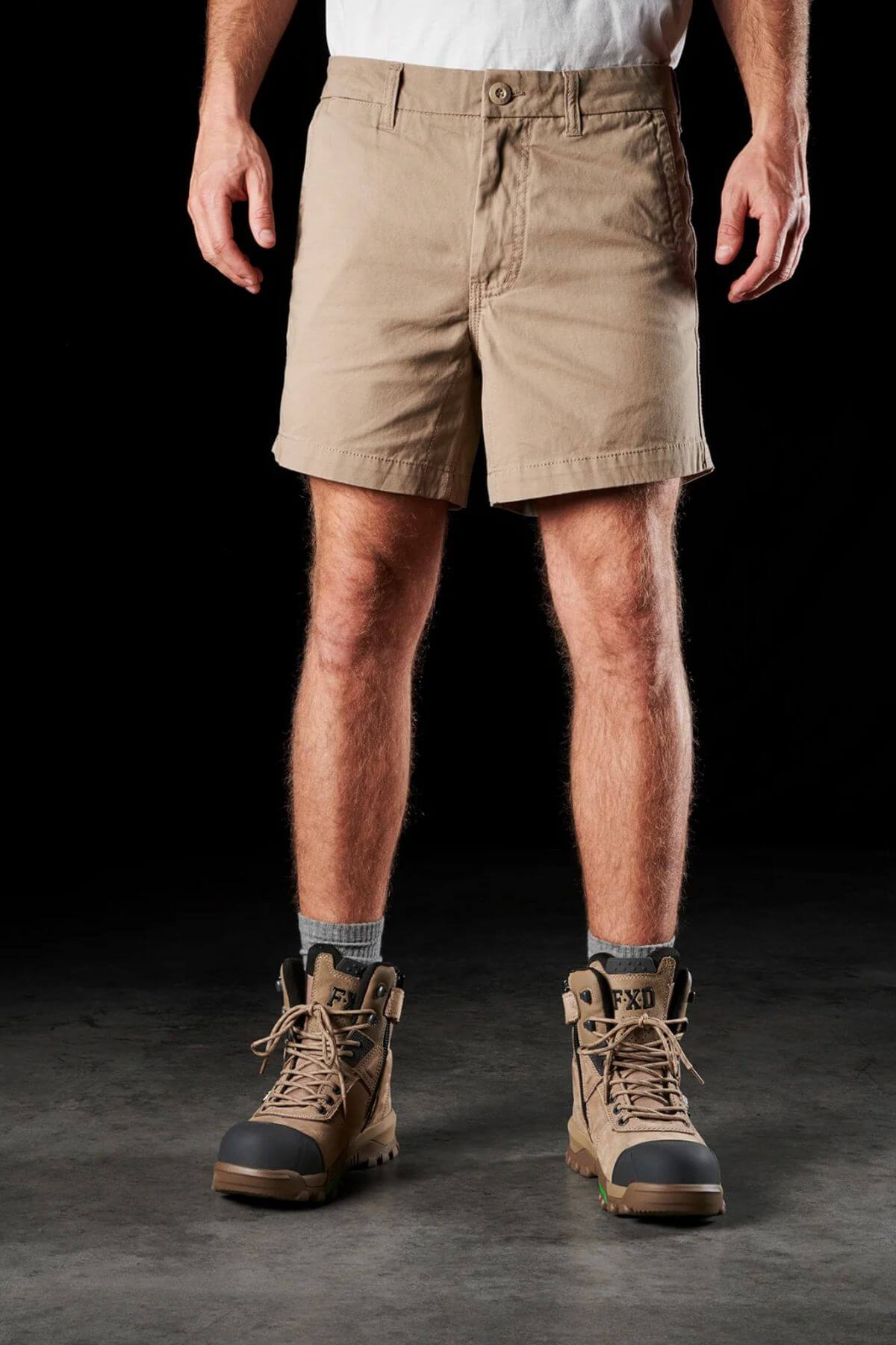 A person is standing on a dark surface, dressed in beige FXD WS-2 Short Shorts made from soft-touch cotton twill, paired with a white shirt, gray socks, and tan lace-up work boots. The image focuses on the lower half of their body against a black background, emphasizing their attire.
