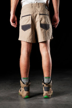 A person wearing the beige FXD WS-2 Short Short, featuring black pocket accents, gray and black socks, and brown hiking boots. The image captures the individual from the back, standing on a concrete floor against a black backdrop.