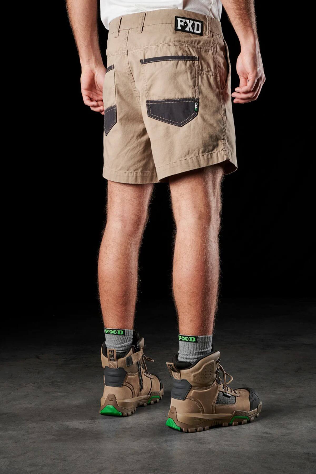 A person is standing, facing away, wearing FXD WS-2 Short Shorts made from soft touch cotton twill with black pockets and an "FXD" logo on the waistband. They are also wearing gray socks and brown work boots with green accents, all set against a black background.