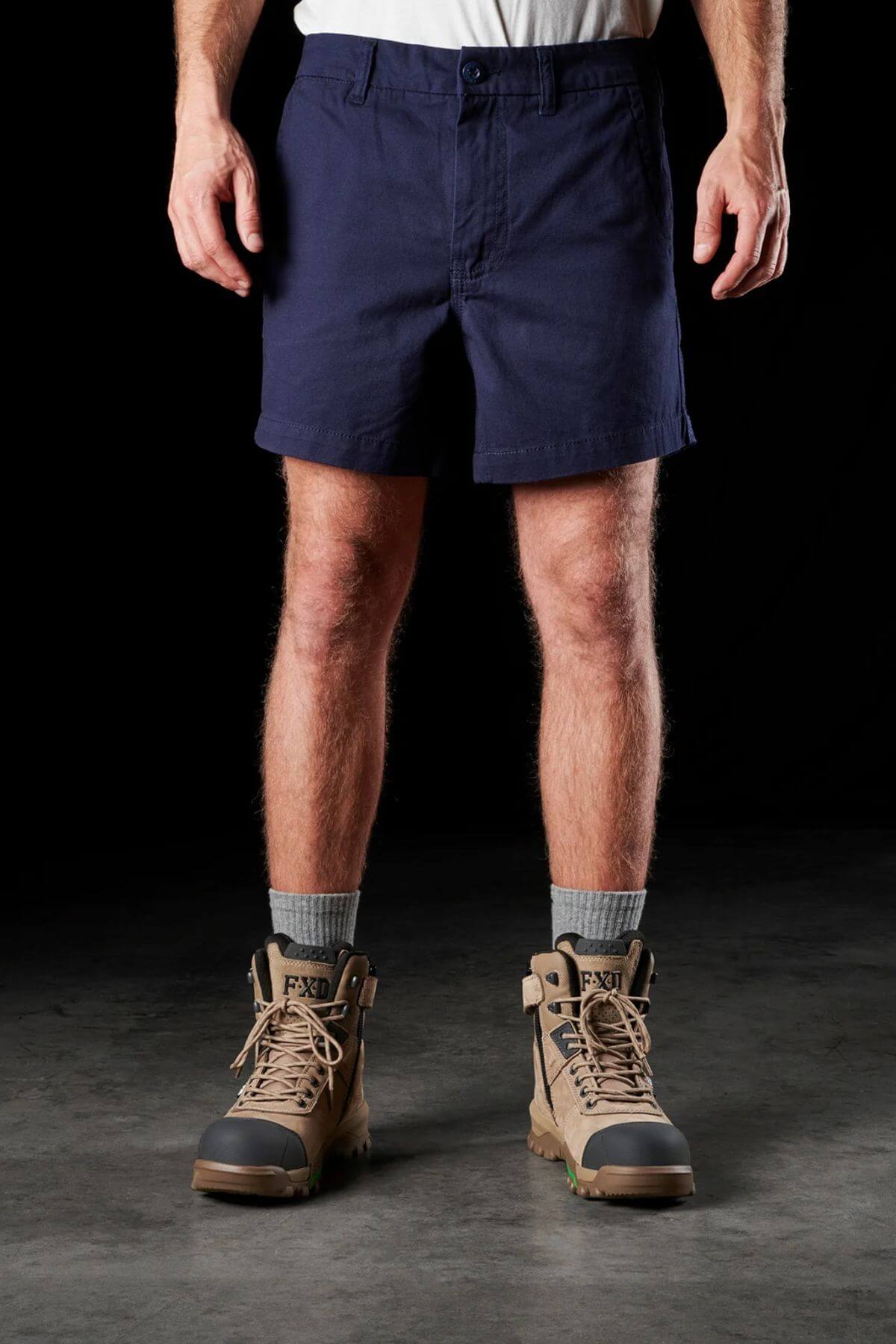 A person wearing FXD WS-2 Short Shorts in durable dark blue, a white shirt, gray socks, and tan work boots stands against a black background. Only the lower part of the body is visible in the image.