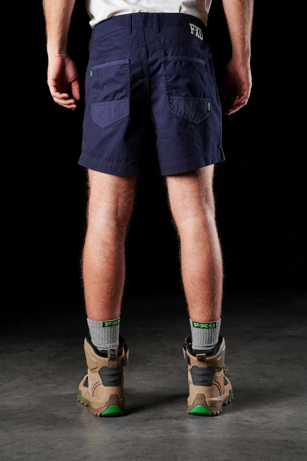 A person is standing with their back to the camera, dressed in FXD WS-2 Short Shorts, which are durable and dark blue with multiple pockets. They have paired the shorts with gray socks and brown hiking boots featuring green soles. The background is dark and indistinct.