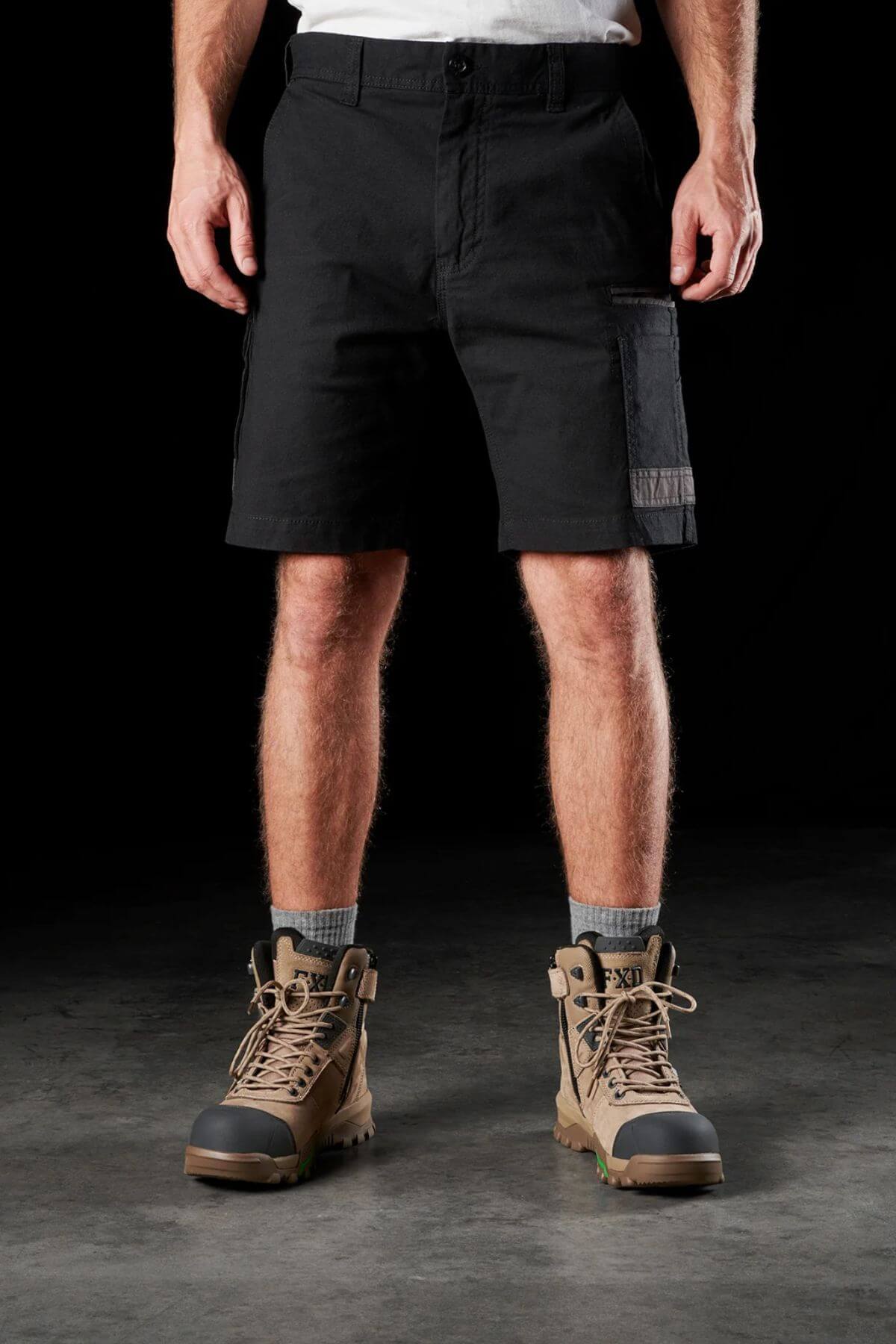 A person stands against a black background wearing FXD WS-3 Shorts made from stretch cotton canvas, gray socks, and tan work boots with black soles. The person's hands rest by their sides, and only their lower body is visible.