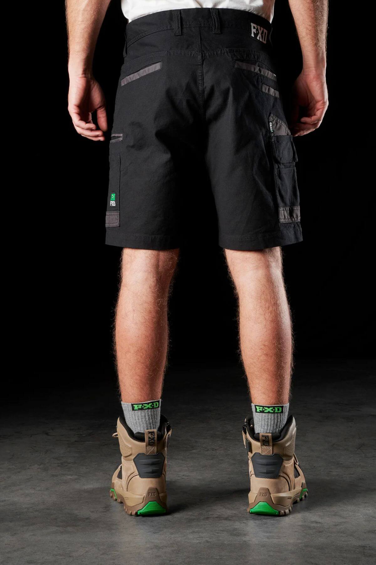 A person stands facing away, wearing FXD WS-3 Shorts made from black stretch cotton canvas with multiple pockets and a white waistband. The durable Dura500™ construction and triple needle seams ensure longevity. They pair these shorts with brown hiking boots featuring green accents and gray socks adorned with "FXD" on the top. The background is dark.