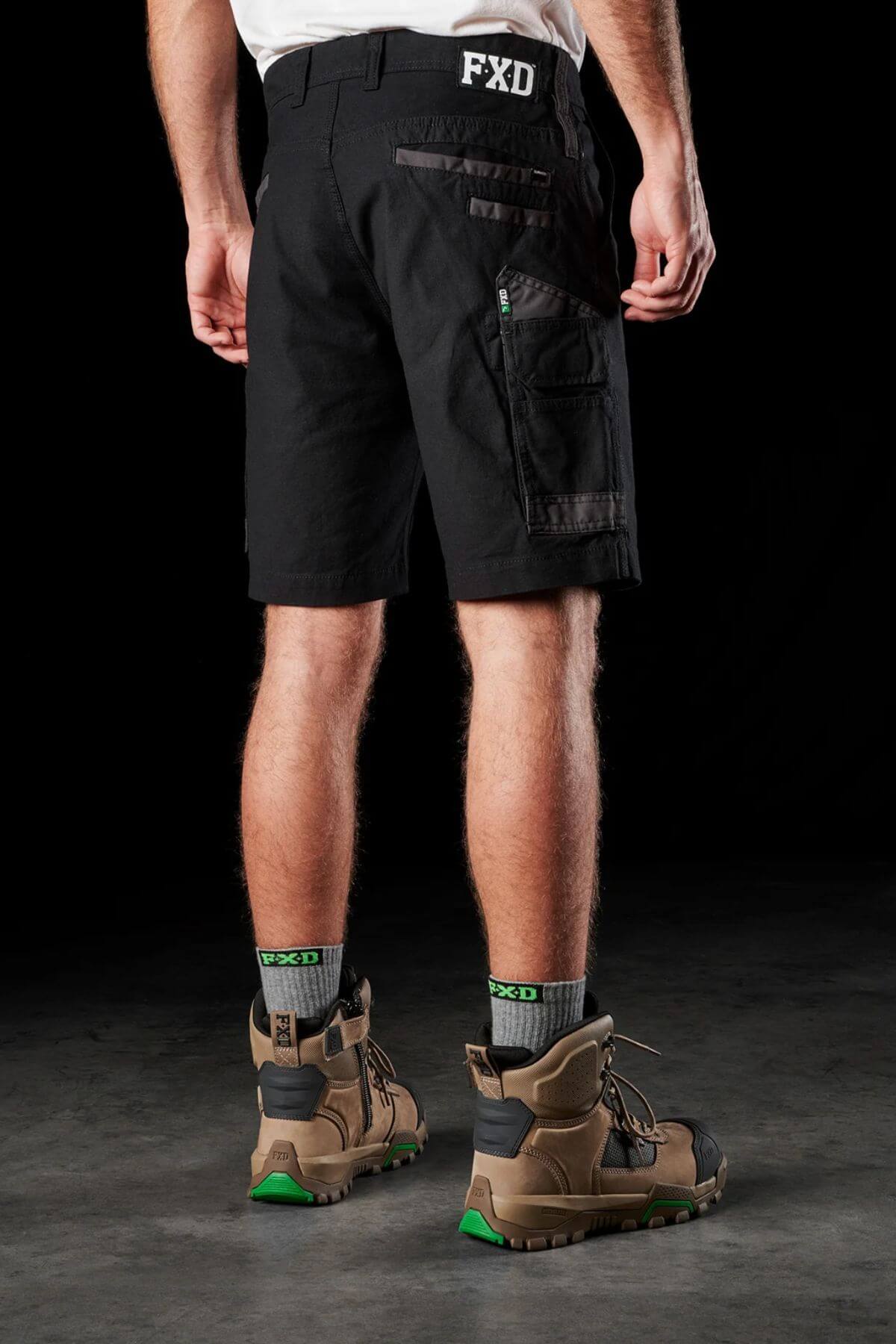 A person is standing with their back facing the camera, wearing FXD WS-3 black cargo shorts with multiple pockets, crafted from stretch cotton canvas with triple needle seams. They are also wearing green and black FXD branded socks and beige and black work boots. The dark background highlights the durable nature of the Dura500™ clothing and footwear.