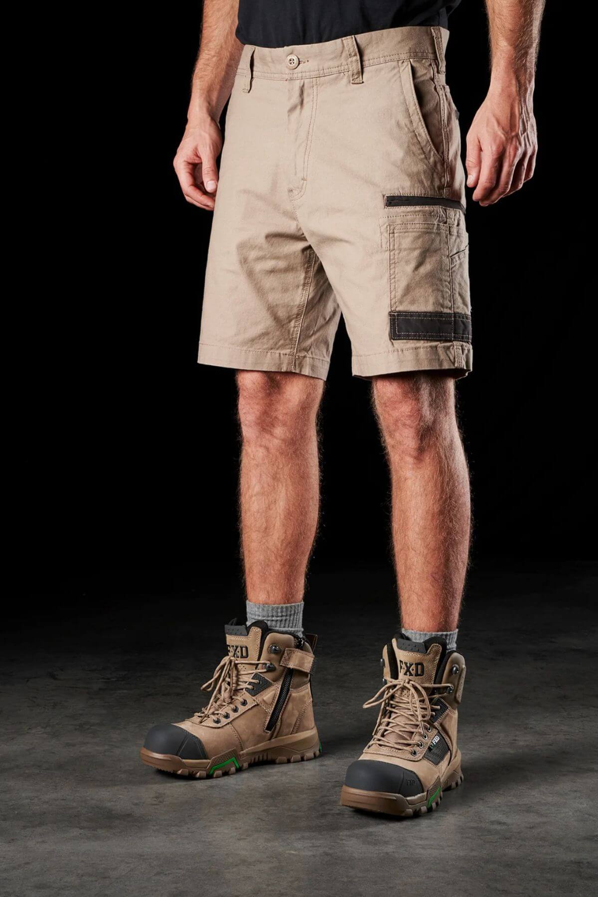 A person wearing beige FXD WS-3 Shorts, a black shirt, and rugged tan work boots with green soles stands against a dark background. Their hands are not visible as they are out of the frame. The FXD WS-3 Short, made from stretch cotton canvas, features multiple pockets and triple needle seams and is slightly wrinkled.