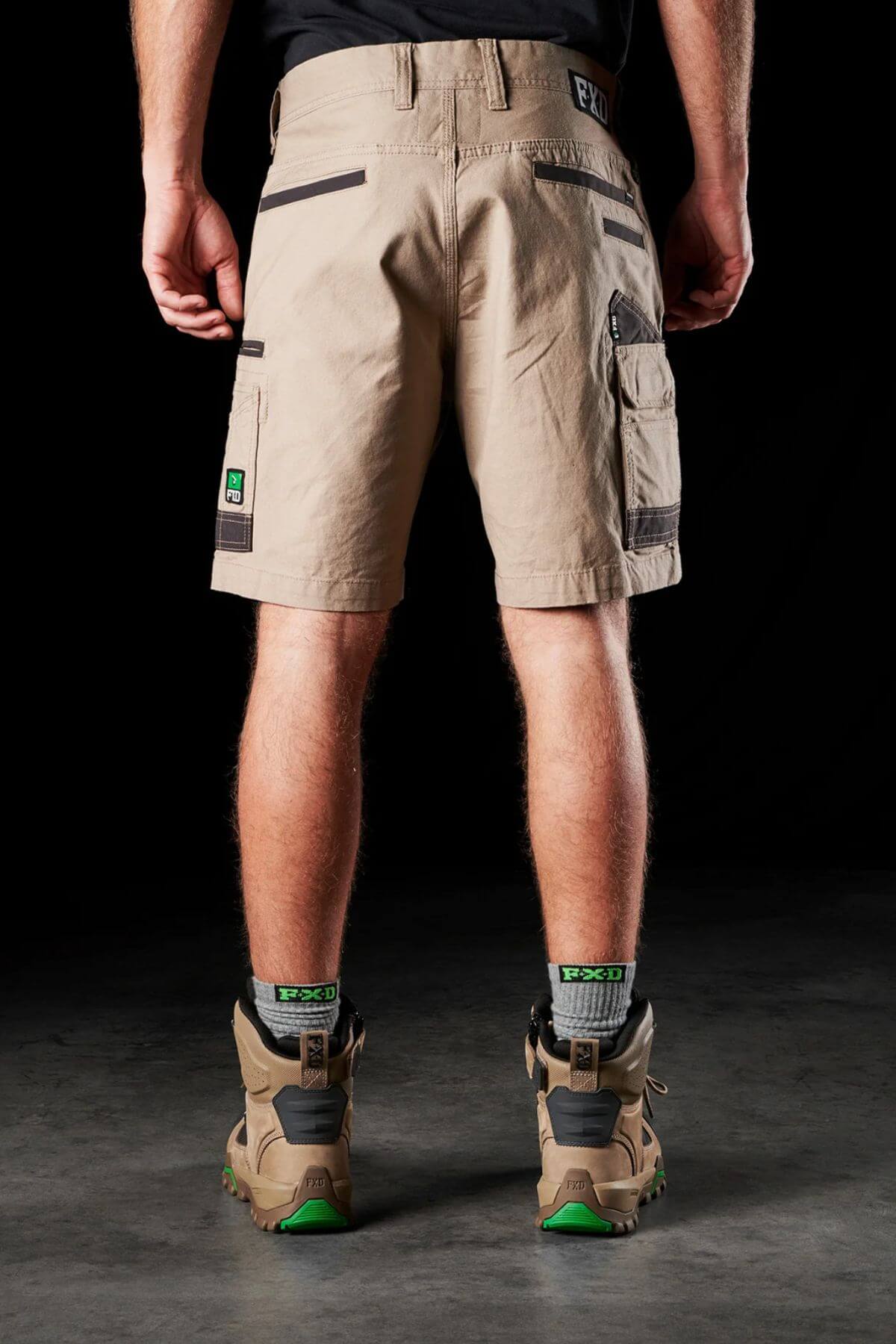 Rear view of a person wearing stretch cotton canvas FXD WS-3 Shorts in beige, along with hiking boots and black-green socks. The background is black, and the person's arms are relaxed by their sides.
