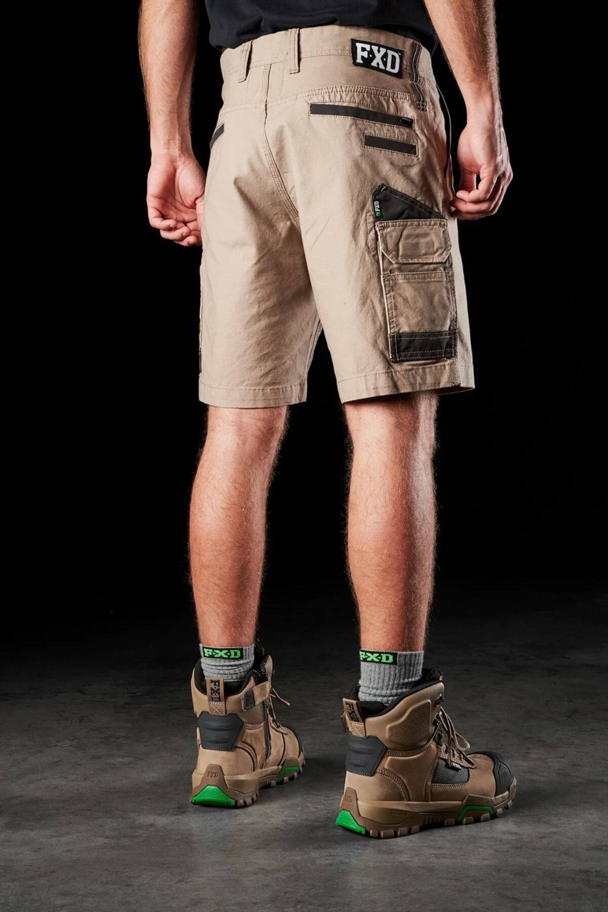A person stands with their back to the camera, wearing beige FXD WS-3 Shorts made from stretch cotton canvas and high-top brown and black work boots. The shorts feature multiple pockets, including one secured with a zipper and triple needle seams. They are also wearing black socks with green, white, and gray lettering.