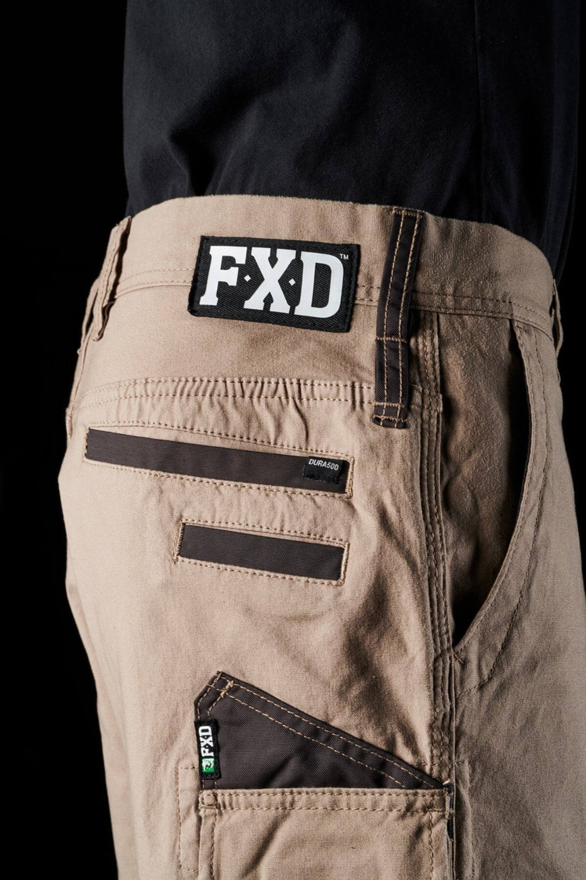 Close-up of a person wearing FXD WS-3 Shorts with brown stitching and multiple pockets. The shorts, crafted from stretch cotton canvas, feature a tag with the logo "FXD" on the waistband and another smaller tag that reads “DURA500” on a pocket. Triple needle seams add durability. The background is black.