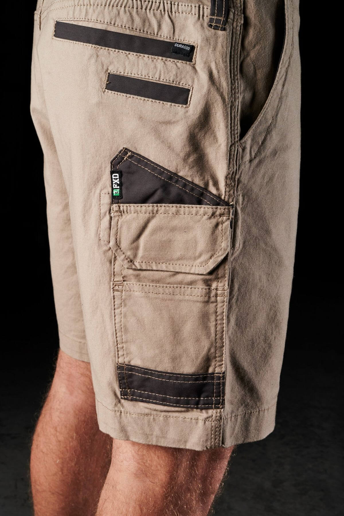 Close-up of a person's legs wearing the FXD WS-3 Shorts in beige, featuring black trim details and multiple utility pockets. Crafted from stretch cotton canvas for durability, these shorts sport a rugged look with reinforced triple needle seams and include a small pocket labeled "CAT." The background is black and indistinct.