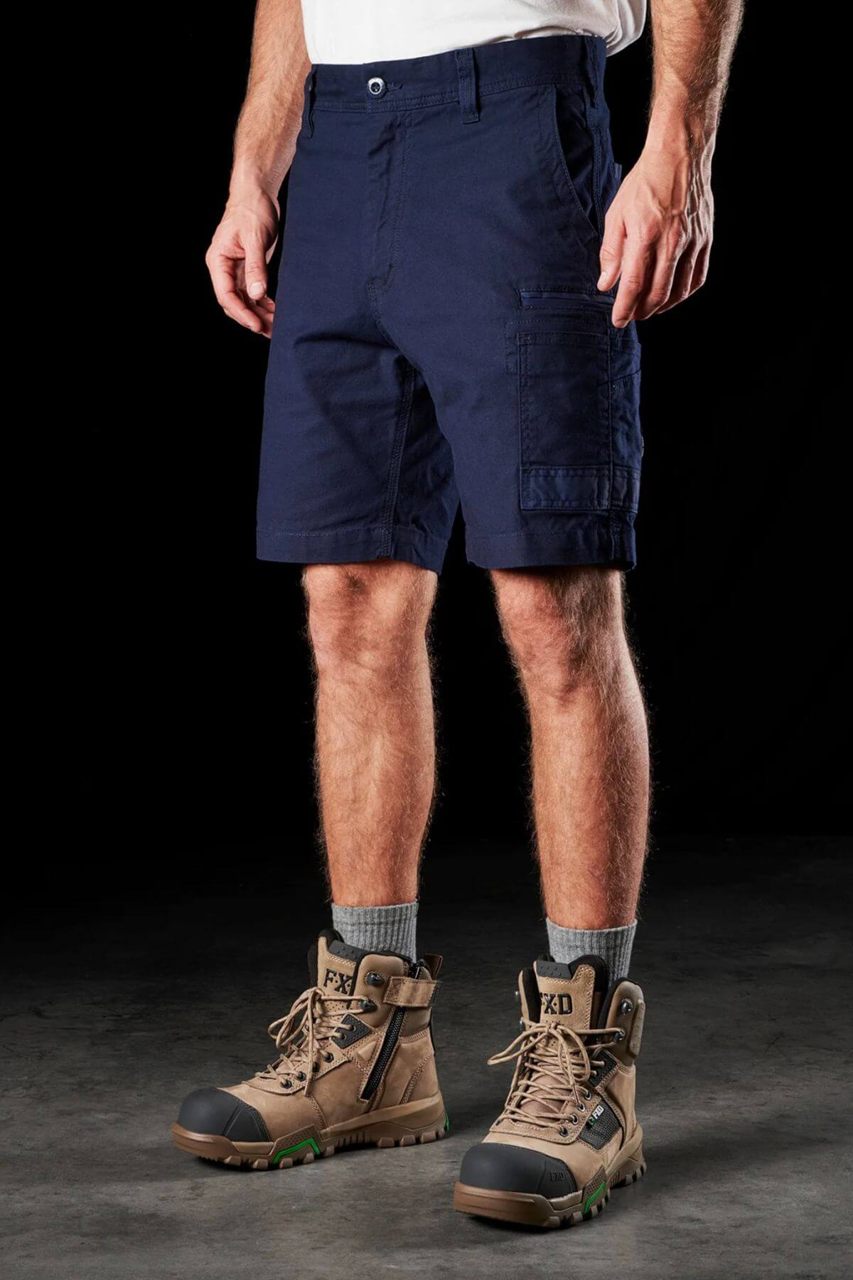 A person wearing FXD WS-3 Short in navy blue, made from stretch cotton canvas, grey socks, and tan high-top work boots stands on a dark surface. The shorts, designed with multiple pockets for added convenience, pair well with the boots that feature a black toe cap and green accents. The outfit exemplifies comfort and durability.