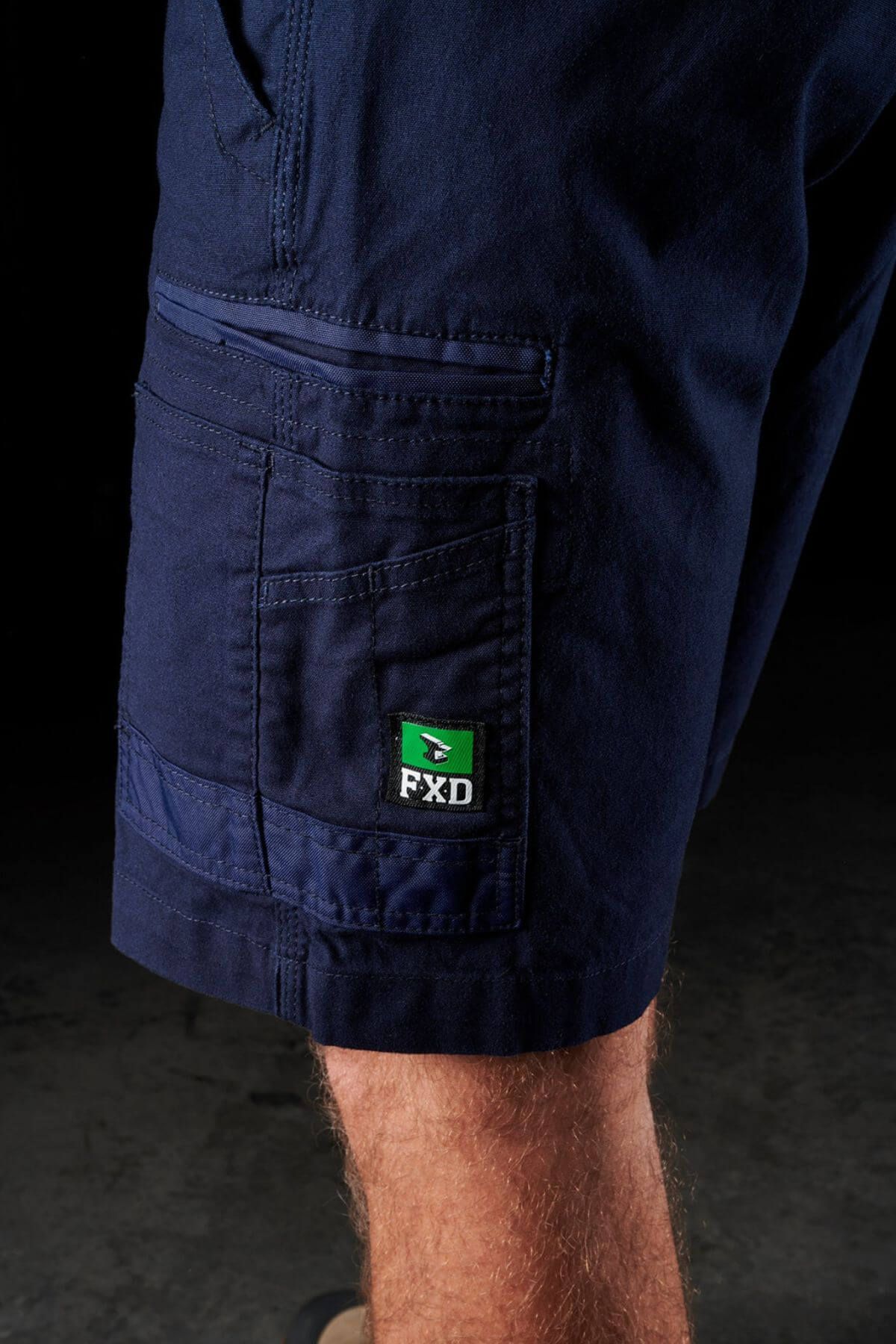 Close-up of a person wearing the dark blue FXD WS-3 Short made from stretch cotton canvas. The image focuses on the lower leg and side pocket area, showcasing detailed stitching and a visible green and black FXD logo tag on the pocket. The background is dark and blurred, emphasizing the resilience of these shorts.