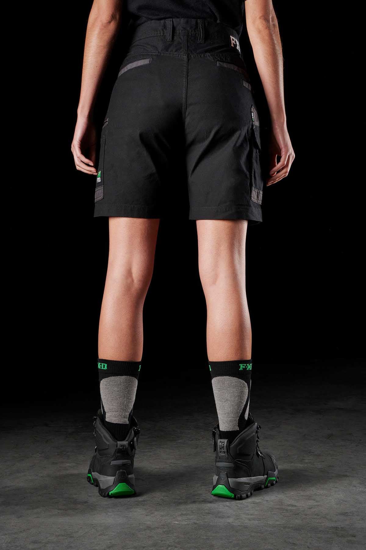 A person stands facing away from the camera, wearing FXD Womens WS-3W Shorts with DURA500 reinforced pockets, gray and black socks, and black boots with green accents. The background is dark, and the legs are partially visible from the calves down to the feet.