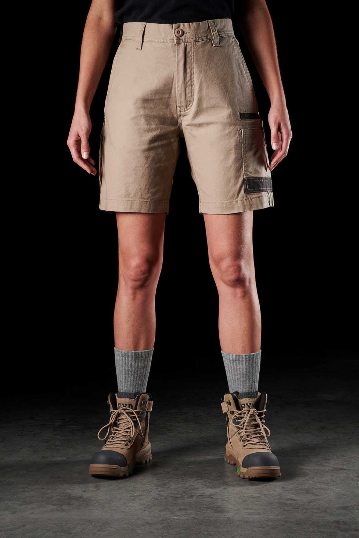 A person is standing, wearing FXD Womens WS-3W Shorts in beige with DURA500 reinforced pockets, a black t-shirt, grey socks, and brown hiking boots. The shorts feature multiple pockets, including one with a black strap. The background is entirely black, and the person is visible from shoulders to boots.