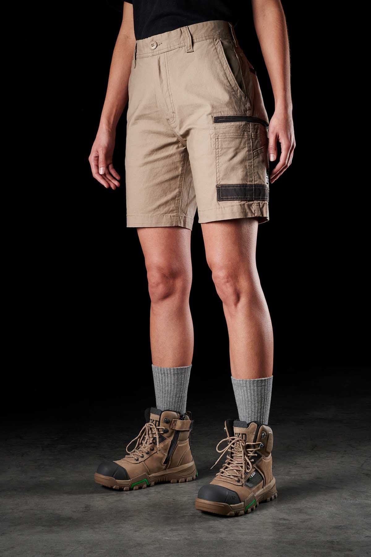 A person stands wearing FXD Women's WS-3W Shorts in beige with DURA500 reinforced pockets, paired with a black belt, gray calf-length socks, and tan hiking boots featuring green accents. The dark background sets off the clothing and footwear.