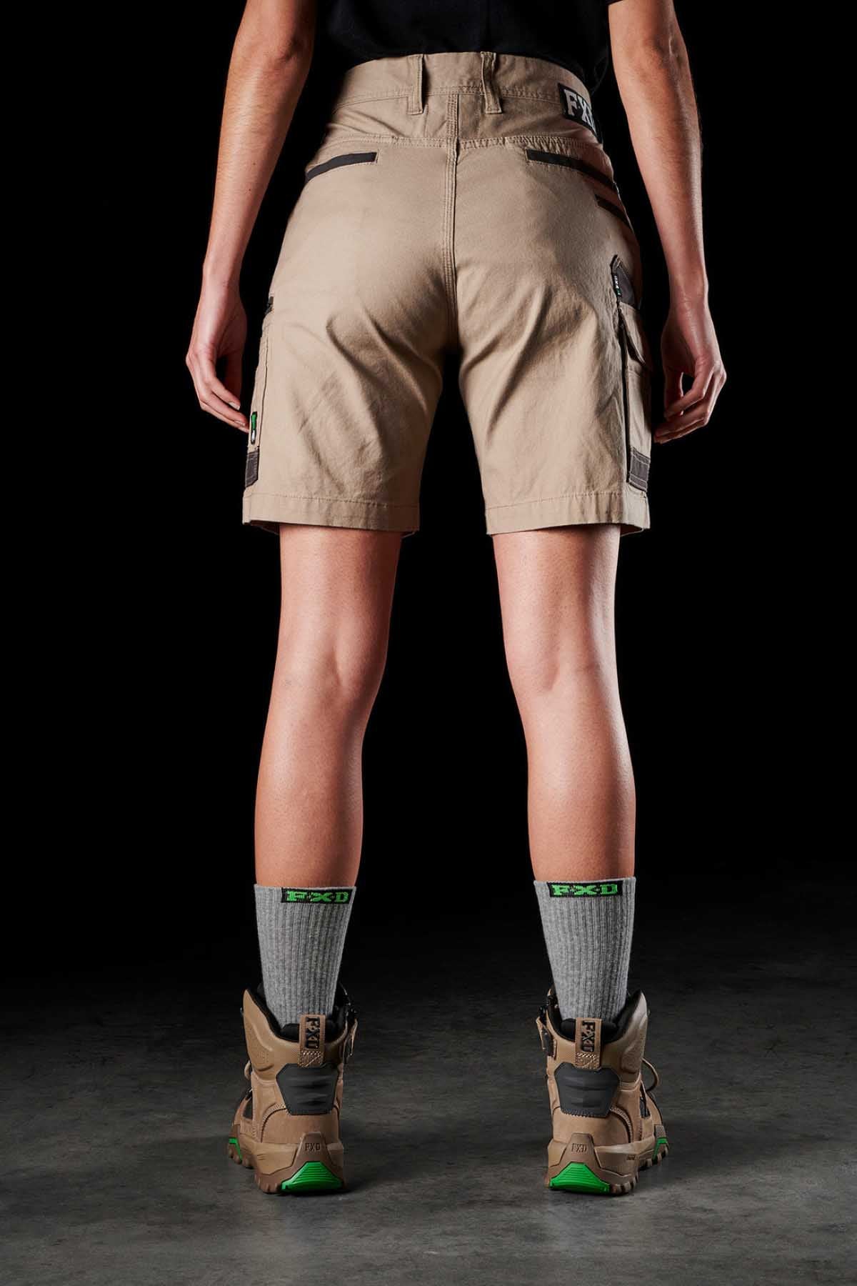 A person stands facing away, wearing FXD's Womens WS-3W khaki utility shorts with DURA500 reinforced pockets, a black top, gray socks with green patterns, and brown hiking boots with bright green soles. The background is a dark, neutral setting.
