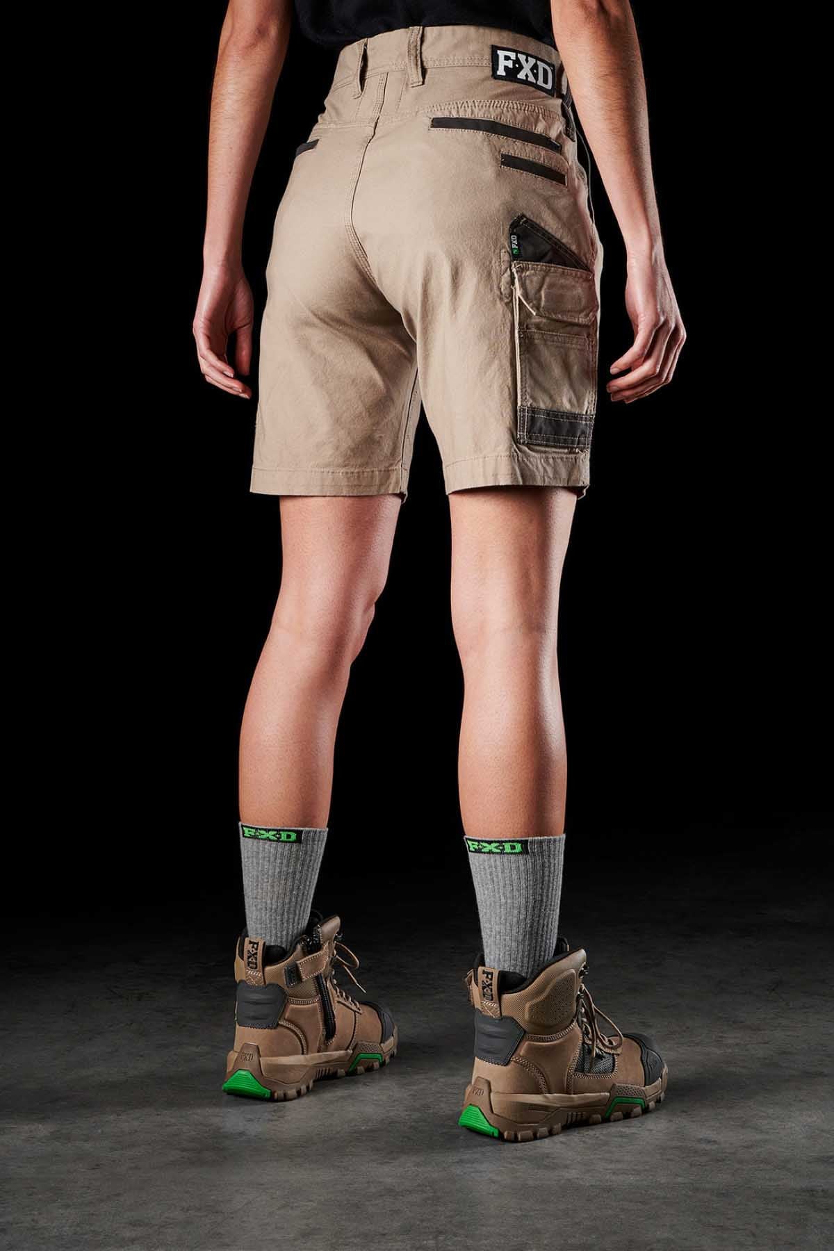 A person stands with their back to the camera, wearing FXD Womens WS-3W khaki stretch work shorts featuring DURA500 reinforced pockets, gray socks, and brown work boots with green accents. The background is plain and dark, emphasizing the clothing and footwear.