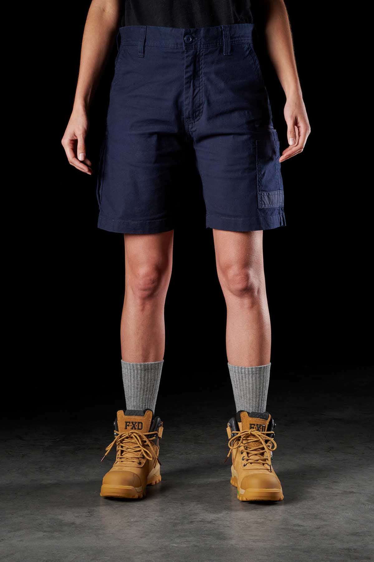A person is standing wearing FXD Womens WS-3W Short in navy blue with DURA500 reinforced pockets, a black shirt, gray socks, and tan work boots. The background is black, and the focus is on the lower half of the body, showcasing the shorts and boots with triple needle seams for added durability.
