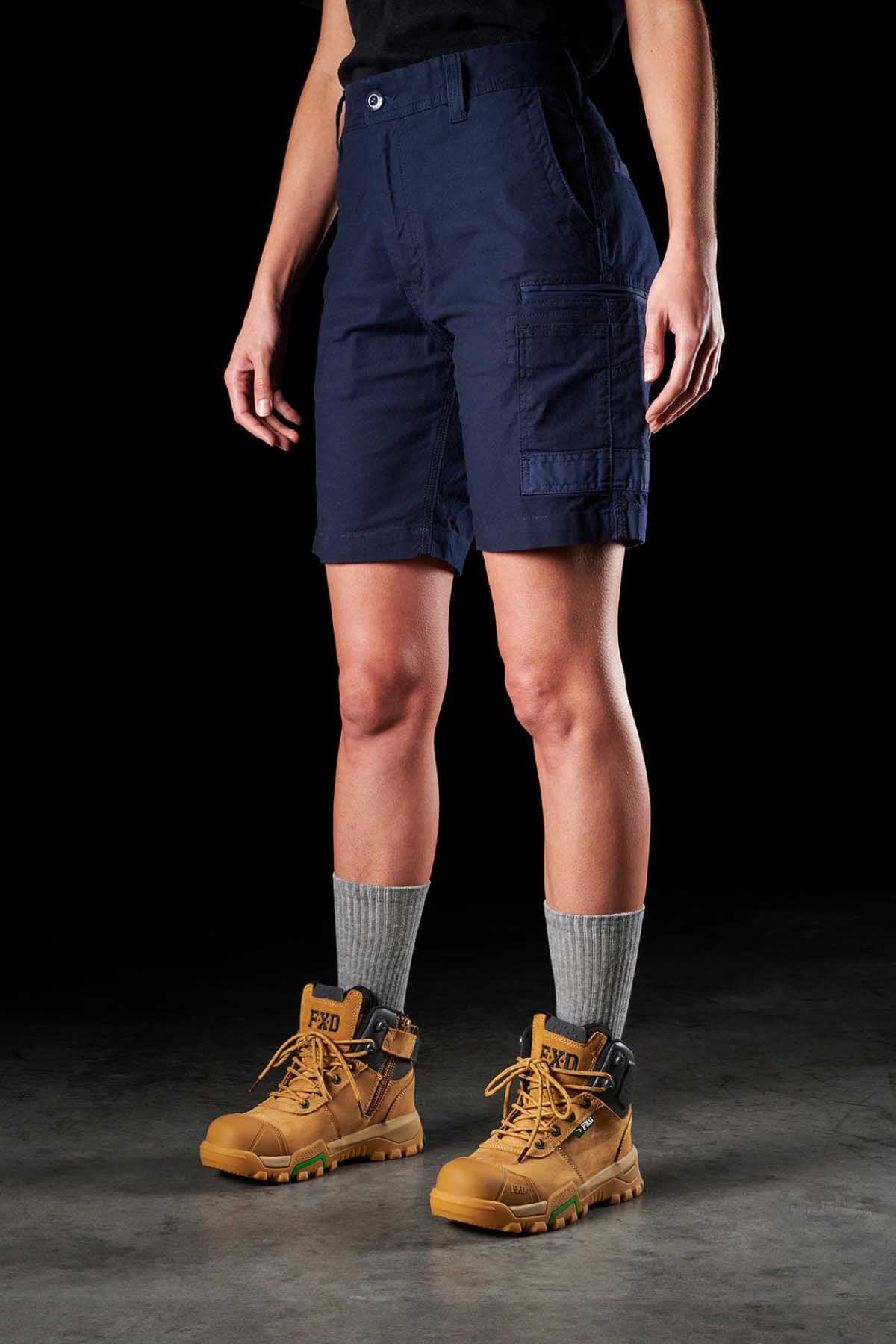 A person is wearing FXD Womens WS-3W Short in navy with triple needle seams, accompanied by gray ribbed socks and tan work boots featuring the "FXD" branding on the tongue and heel tabs. Their hands rest at their sides against a black background.