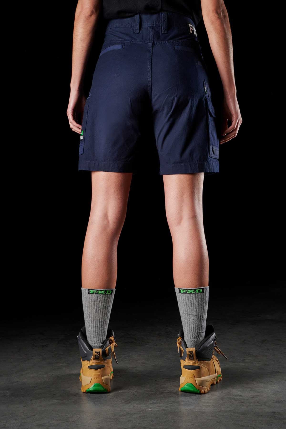 A person stands with their back to the camera, wearing the FXD Womens WS-3W Short in dark blue stretch fabric, featuring DURA500 reinforced pockets. They are also sporting a black shirt, grey socks, and tan hiking boots. The dark background enhances the contrast of their sturdy attire and footwear.