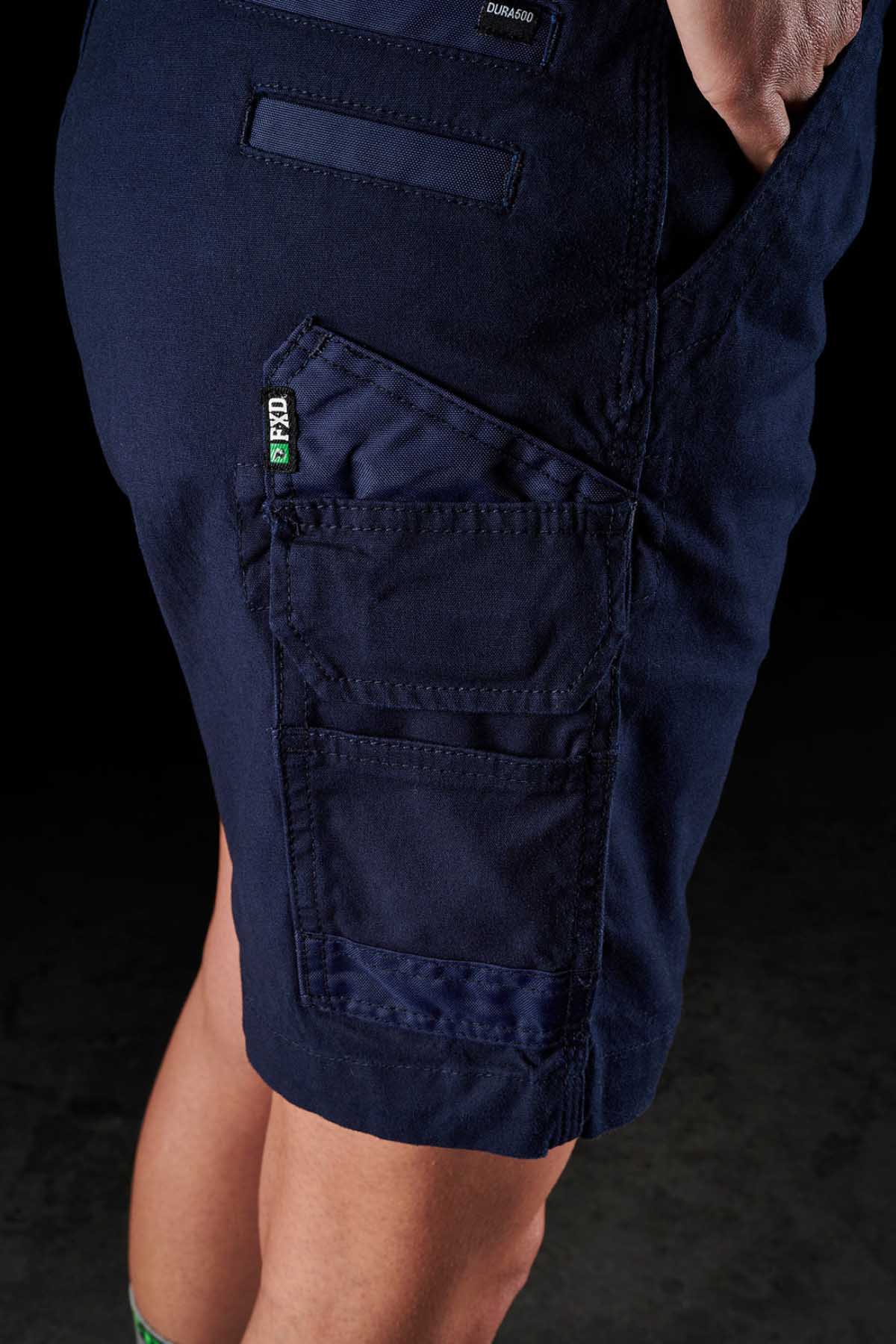 Close-up of a person wearing FXD Womens WS-3W Shorts in navy blue, featuring triple needle seams and multiple pockets, including a prominent one with a green FPPO tag. The background is dark, and only the lower torso and upper legs of the person are visible.