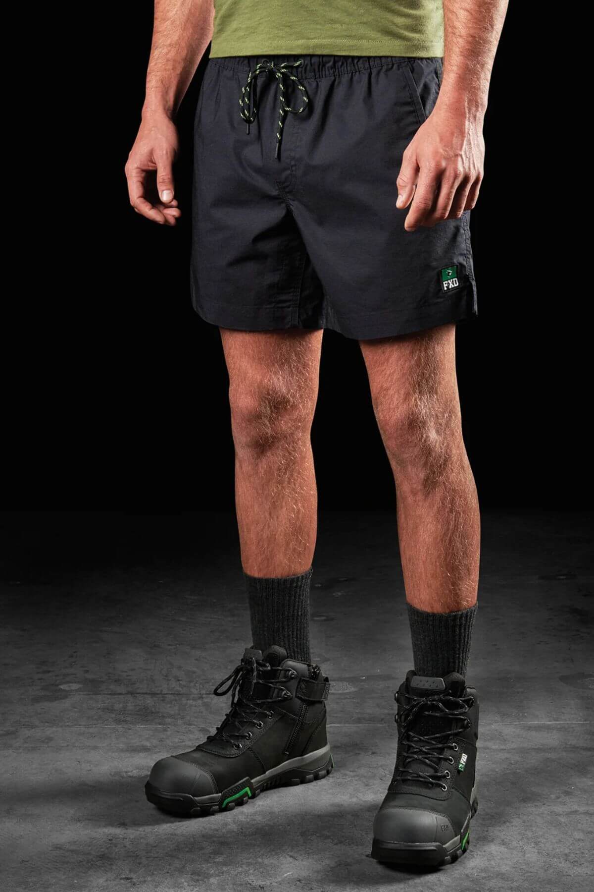 A person stands against a dark background wearing an olive green T-shirt, black socks, rugged black and green hiking boots, and FXD WS-4 Shorts with a comfort stretch elastic waistband and triple needle seams. Only the person's lower torso and legs are visible.