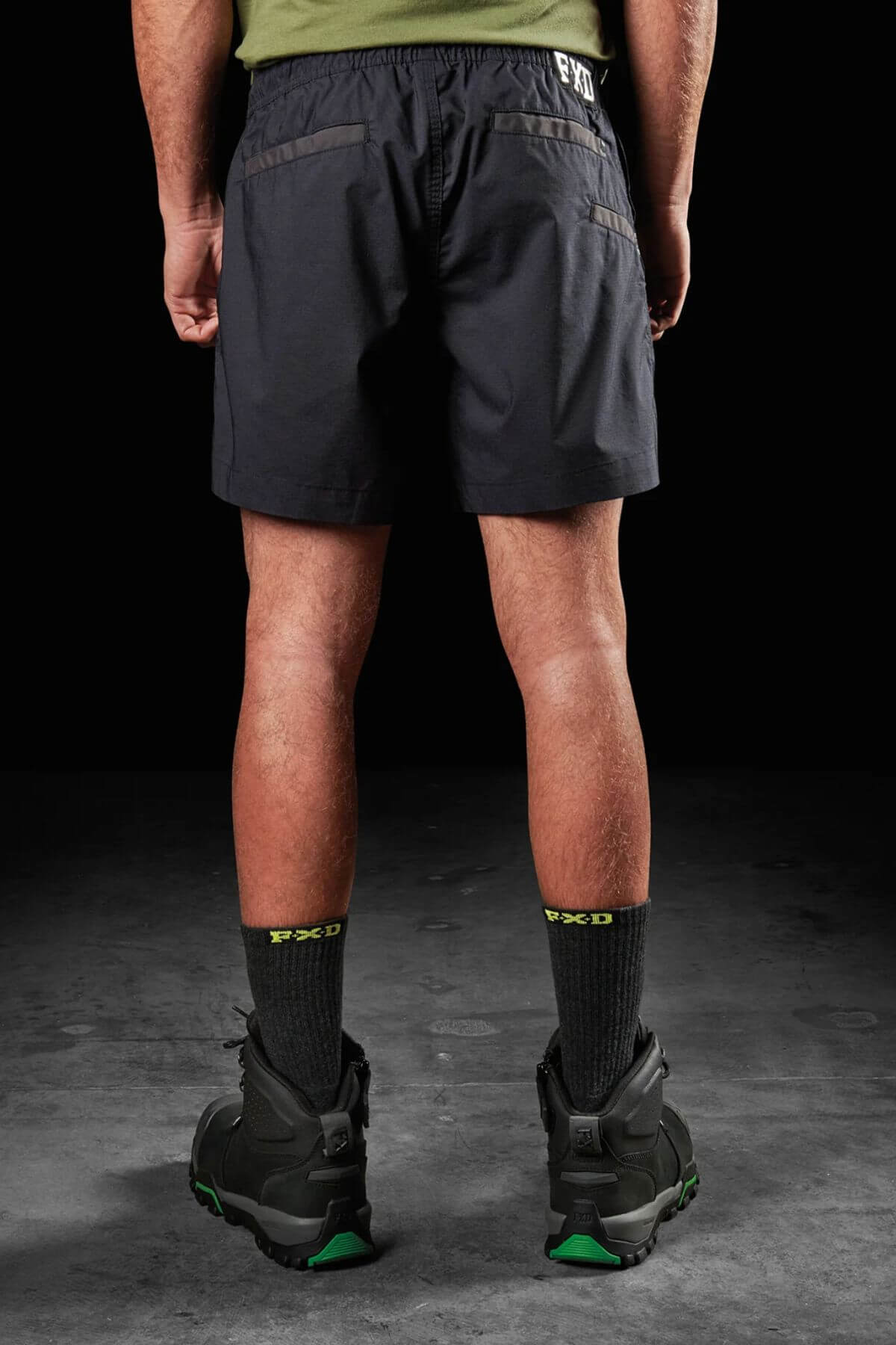 Rear view of a person wearing FXD WS-4 shorts in black, made from recycled polyester stretch ripstop, accompanied by a green shirt and black socks with green lettering. They are standing on a concrete surface and sporting black shoes with green accents. The background is black.