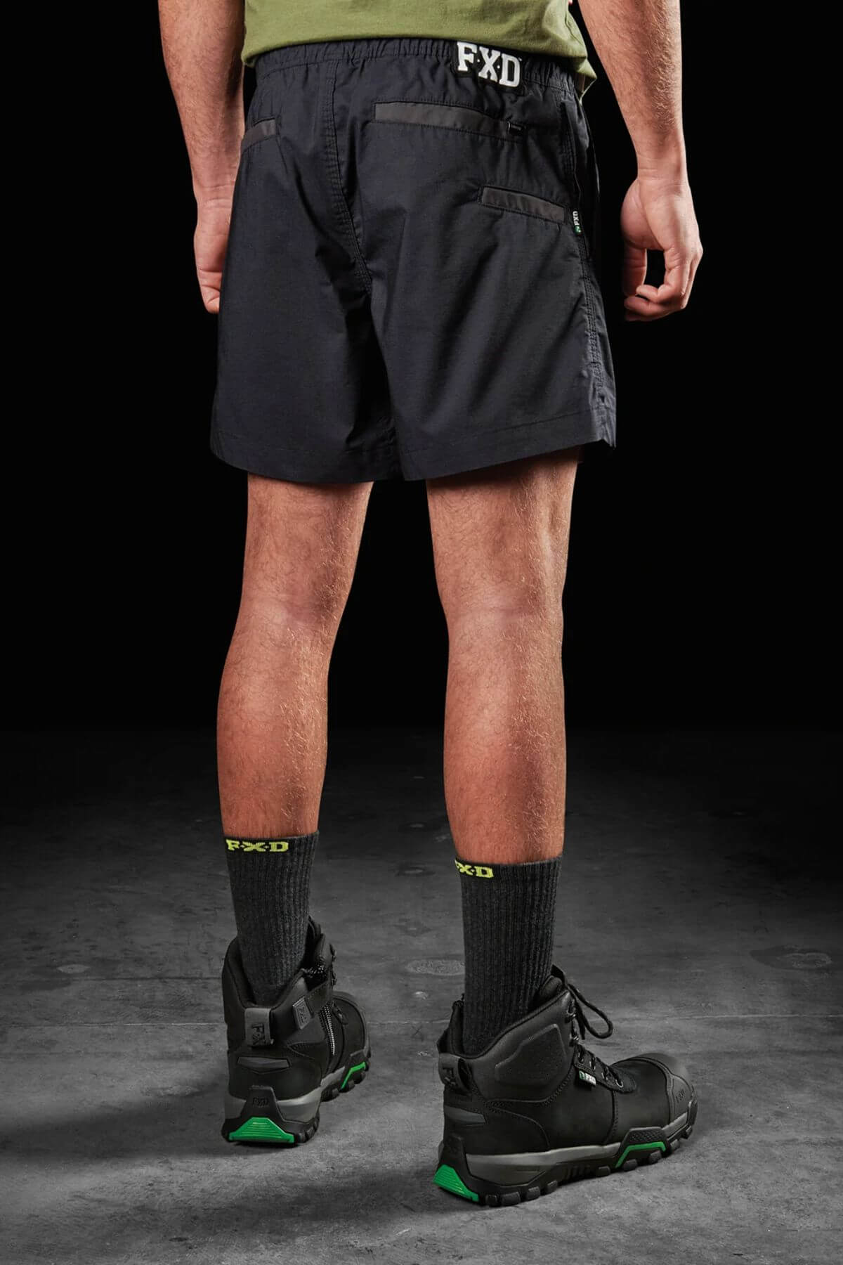 A person stands facing away, wearing a green shirt and FXD WS-4 Short—black shorts with the letters "FXD" on the waistband, featuring triple needle seams and made from recycled polyester stretch ripstop fabric. Black socks with "FXD" written in green, along with black work boots featuring green detailing, complete the look. The background is dark.