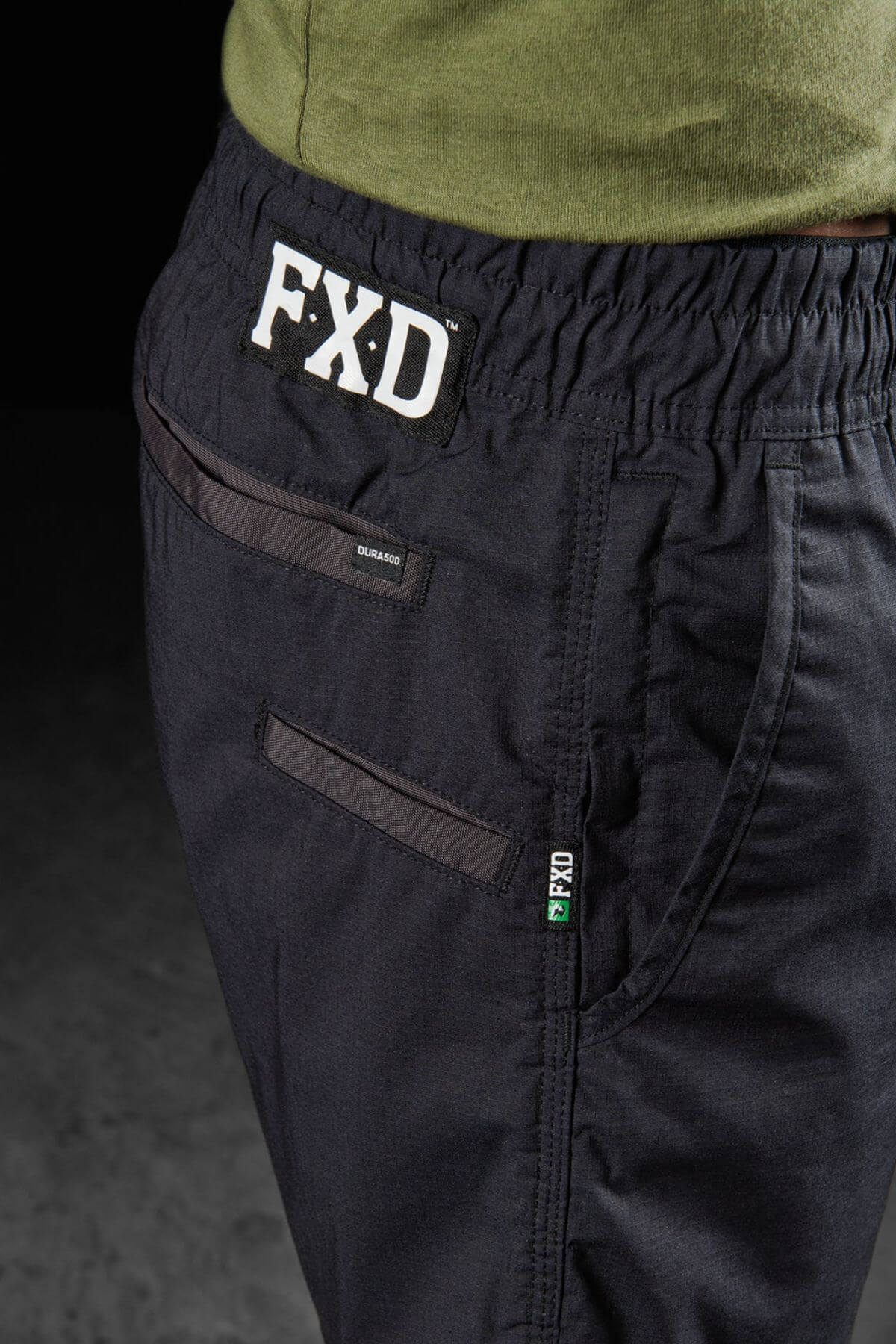 Close-up of a person wearing black FXD WS-4 Shorts made from Recycled Polyester Stretch Ripstop, featuring a comfort stretch elastic waistband with the FXD logo prominently displayed. The shorts have triple needle seams, and one pocket is secured with a zipper while the other with a button, paired with a green shirt tucked in.