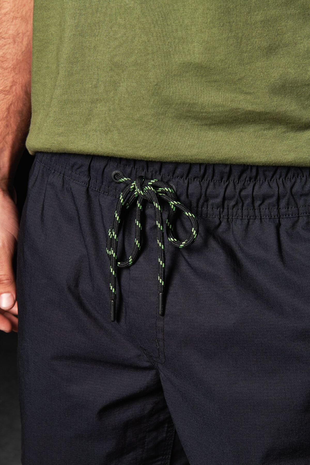 Close-up of a person wearing FXD WS-4 Shorts with a comfort stretch elastic waistband and a green T-shirt. Only the torso and part of the arm are visible. The drawstring on the shorts is green with black and white accents, tied in a bow, showcasing meticulous triple needle seams.