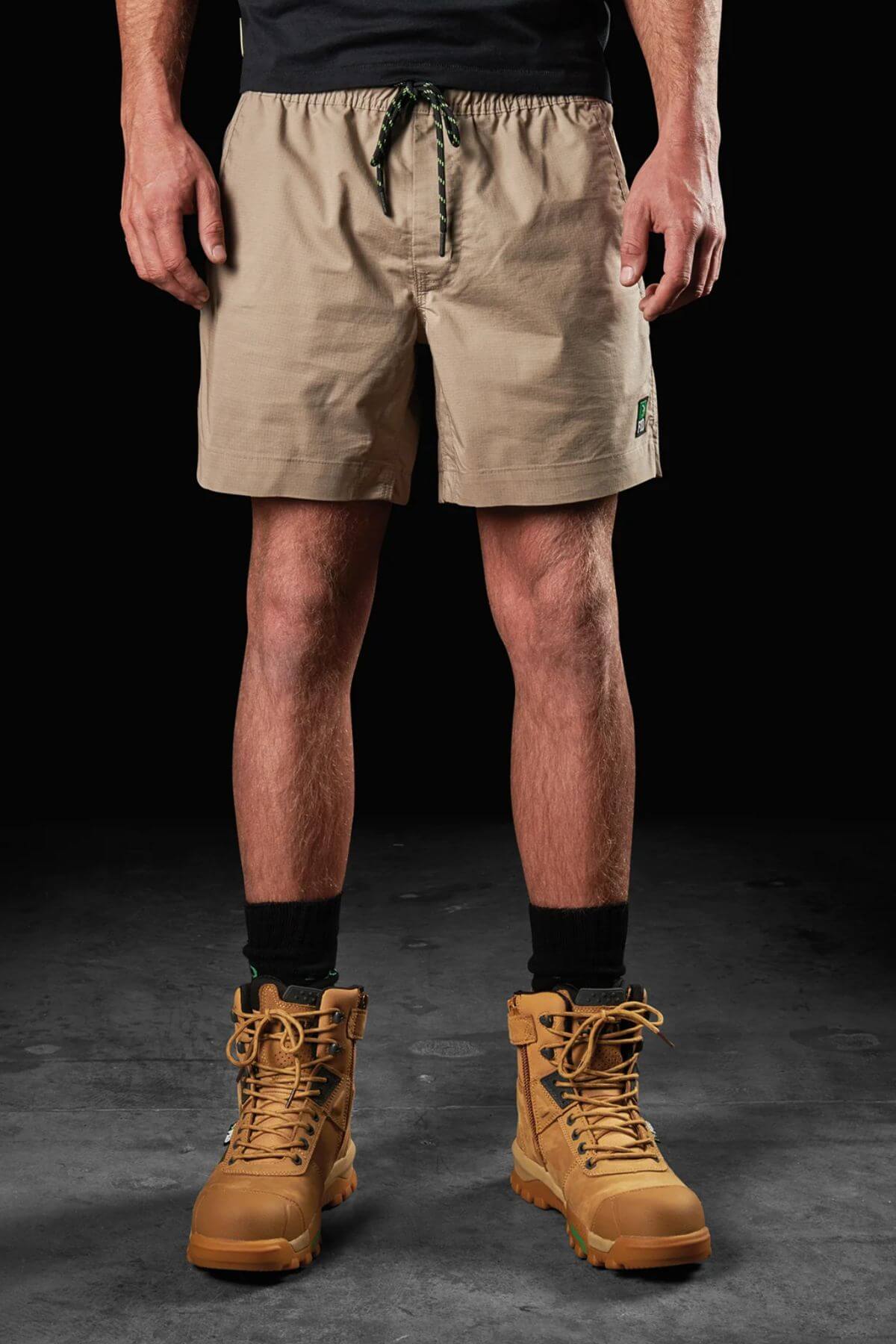 A person stands against a black background wearing FXD WS-4 Shorts in beige, which feature a comfort stretch waistband and drawstring, along with black socks and tan lace-up boots. The individual’s upper body is mostly out of frame, with their hands relaxed by their sides. The dark and textured floor adds depth to the scene.