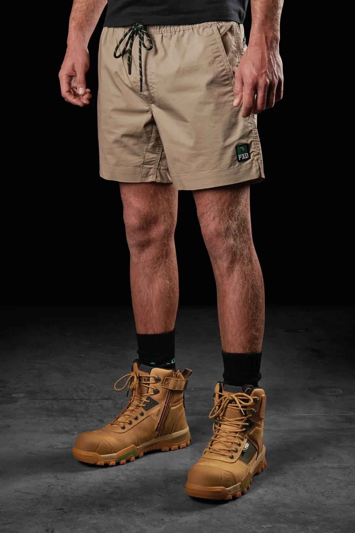 A person is standing against a dark background wearing FXD WS-4 Shorts, which are beige with a small green logo, paired with black socks and light brown lace-up boots. The shorts feature a comfort stretch waistband that ensures ease of movement. Only the lower half of their body is visible.