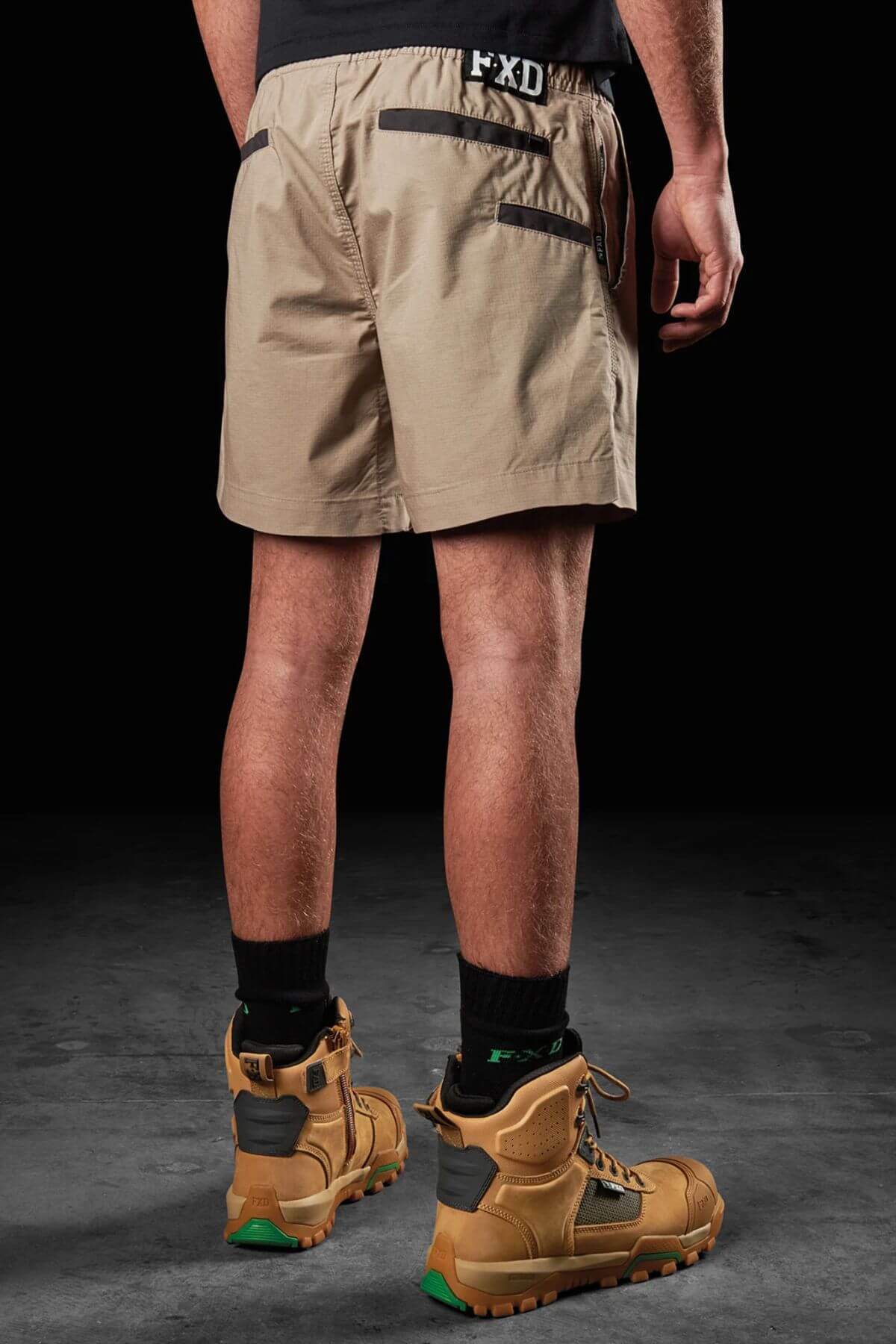 A person stands with their back to the camera, wearing the FXD WS-4 Short featuring a comfort stretch waistband, black socks, and brown work boots on a dark, textured background. The waistband of the shorts displays the letters "F X D" in black and white.