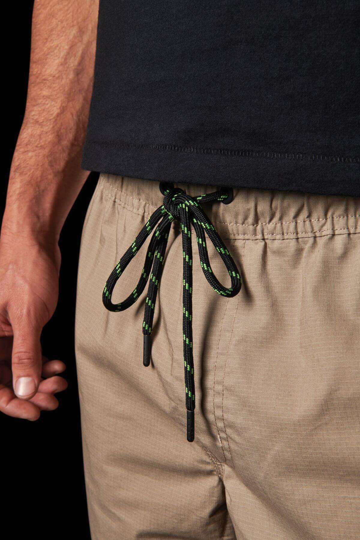 Close-up of the waist of a person wearing FXD WS-4 Shorts in beige, made from Recycled Polyester Stretch Ripstop, featuring a green and black drawstring tied in a bow. The right hand is visible along the side of the shorts with triple needle seams, and the person is wearing a black shirt that slightly overlaps with the waistband.