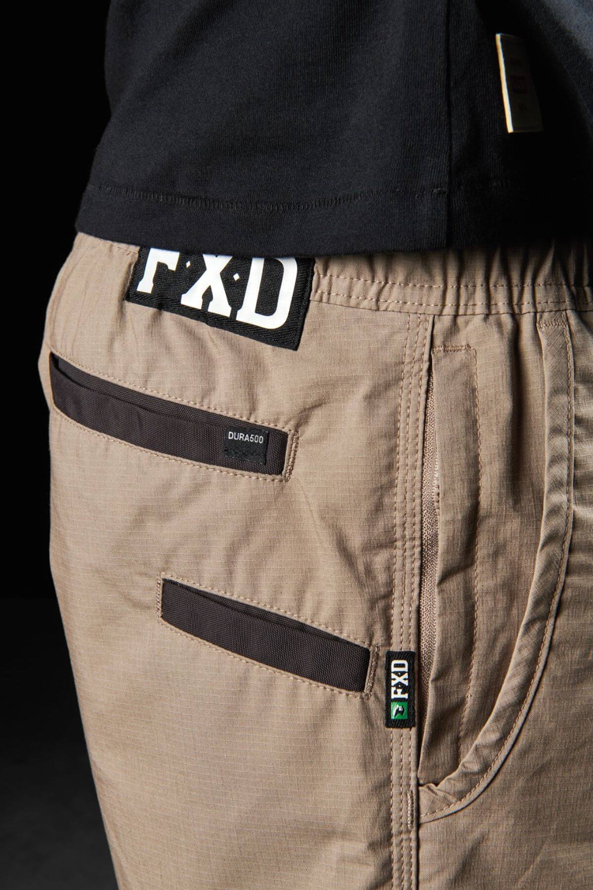 Close-up of a person wearing khaki FXD WS-4 shorts with black trim. The shorts, crafted from recycled polyester stretch ripstop, feature a back pocket with a black label that reads "DURAS60" and another black label on the waistband that reads "FXD." Triple needle seams ensure durability. The person is also wearing a black shirt.