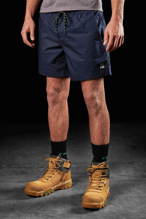 A person is wearing a pair of FXD WS-4 Shorts in dark blue with a comfort stretch waistband, a grey t-shirt, and black socks featuring a green pattern tucked into tan work boots. The background is solid black, and only the person's midsection and legs are visible.