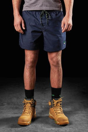 A person stands against a dark background, wearing a pair of FXD WS-4 Shorts in navy blue, featuring a green drawstring and a comfort stretch waistband. A grey shirt is partially visible beneath these shorts. The ensemble is completed with black socks that have green accents and tan work boots. The person's hands rest relaxed by their sides.