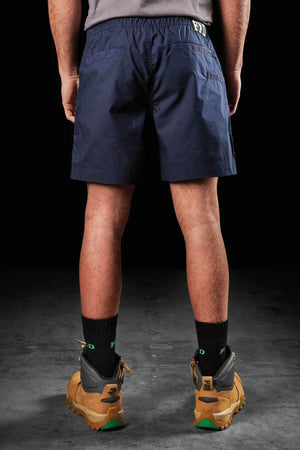 A man stands facing away, wearing black socks, brown work boots, and navy blue FXD WS-4 Shorts featuring the letters "FFD" on the back. These shorts are crafted from organic cotton with a comfort stretch waistband. The background is dark.