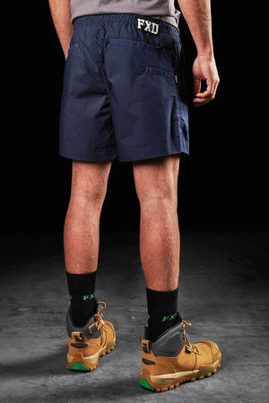 A person is shown from behind wearing dark blue FXD WS-4 Shorts, which feature a comfort stretch waistband, along with black FXD socks and tan lace-up boots accented in green. The dark, unlit background emphasizes the clothing and footwear.