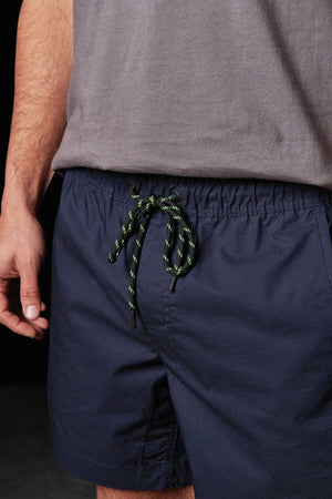 A person wears a gray T-shirt and the FXD WS-4 Short in navy blue, which features an adjustable black drawstring with green accents and a comfort stretch waistband. Their left hand hangs loosely by their side, and the image is cropped to show only their torso and upper legs.