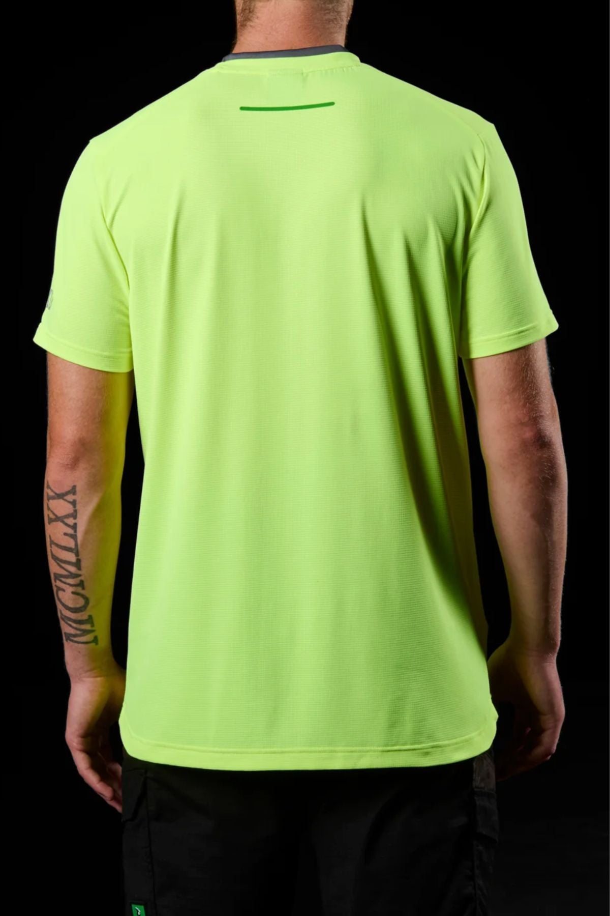 A person is shown from the back against a black background wearing an FXD WT-4 Hi Vis Tech Tee in neon yellow-green. The quick-drying, moisture-wicking shirt highlights a bold tattoo on their right forearm.