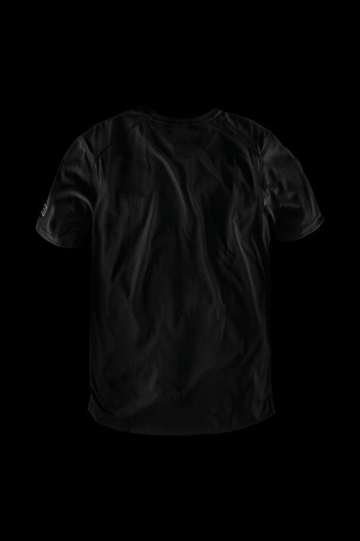 The FXD WT-3 Tech Tee, a black short-sleeve t-shirt renowned for its comfort, is showcased against a solid black background. The shirt features a plain design with subtle wrinkles and a slightly curved hem at the bottom.