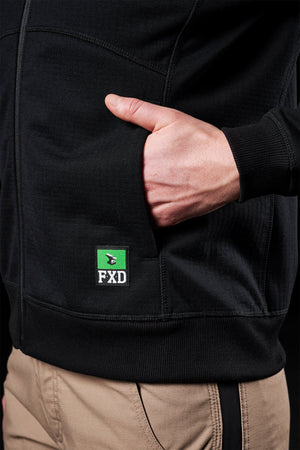 A hand rests comfortably in the pocket of an FXD Women's WF-3W Jacket in black, featuring the logo and windproof qualities, paired with classic tan pants.
