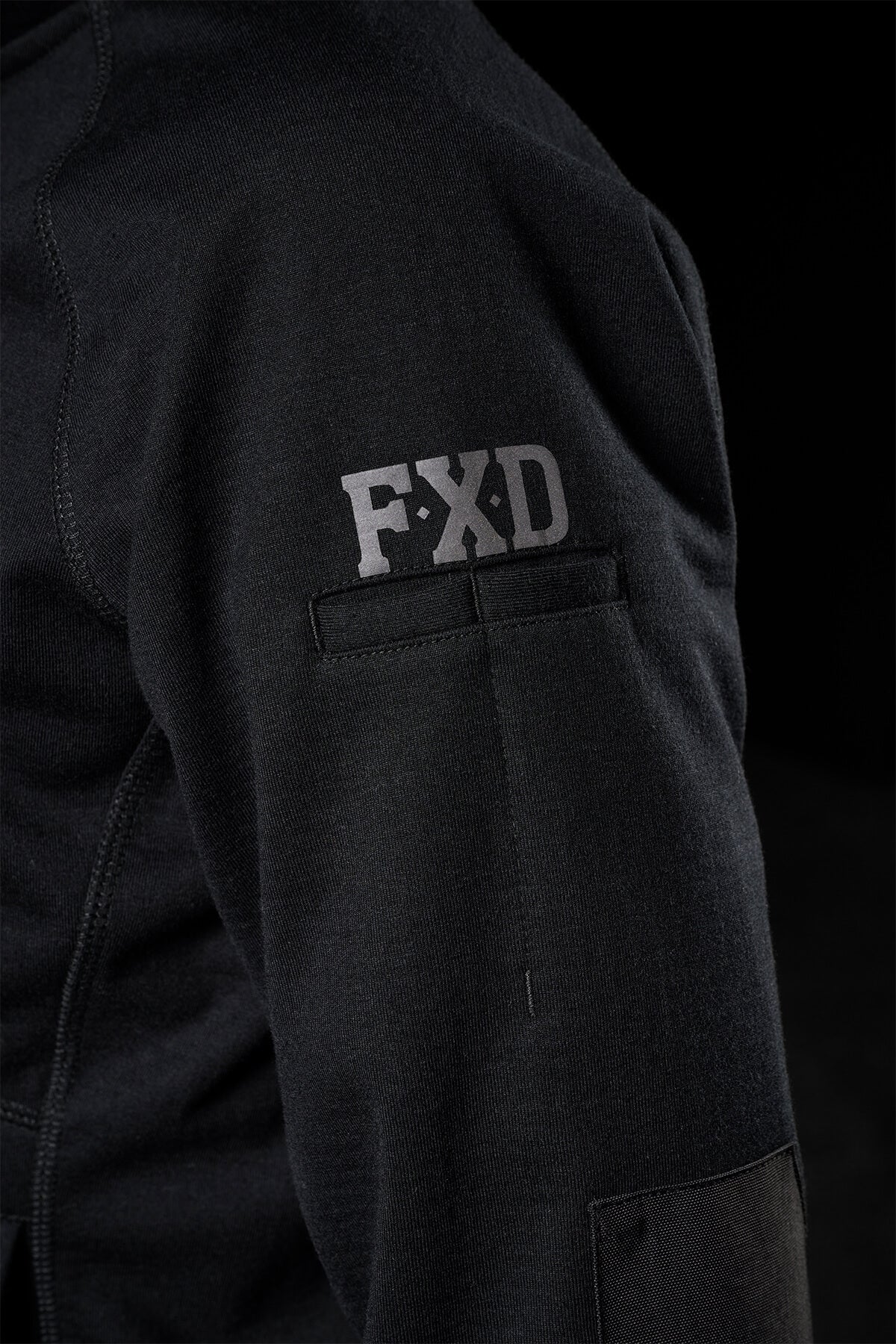 A close-up of the FXD Women's WF-3W Jacket sleeve shows "FXD" near the shoulder. The smooth, water-resistant fabric shines against a dark background, highlighting the texture and detail of this windproof garment.