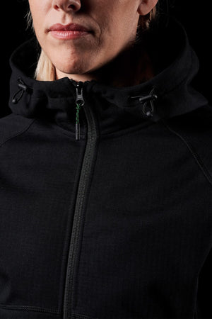 A person dons a black FXD Womens WF-3W Jacket with the hood down against a dark backdrop, highlighting its sleek design. The shot focuses on the upper body and face up to the nose, emphasizing the jacket's windproof fabric and practical drawstrings near the neck.