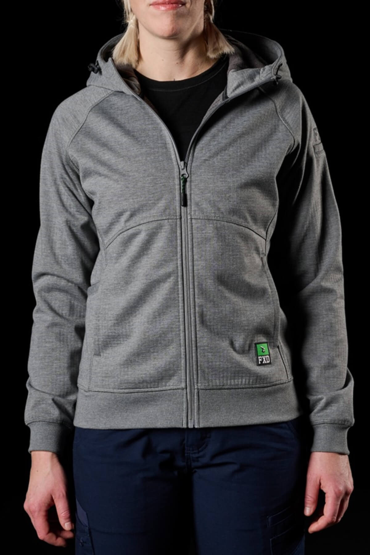A person is standing against a black background, wearing a grey zip-up hoodie with a small green logo on the left pocket. The hoodie, featuring front pockets and a zipper, boasts water resistance and windproof qualities. The person is also wearing dark pants that are reminiscent of the FXD Womens WF-3W Jacket style.