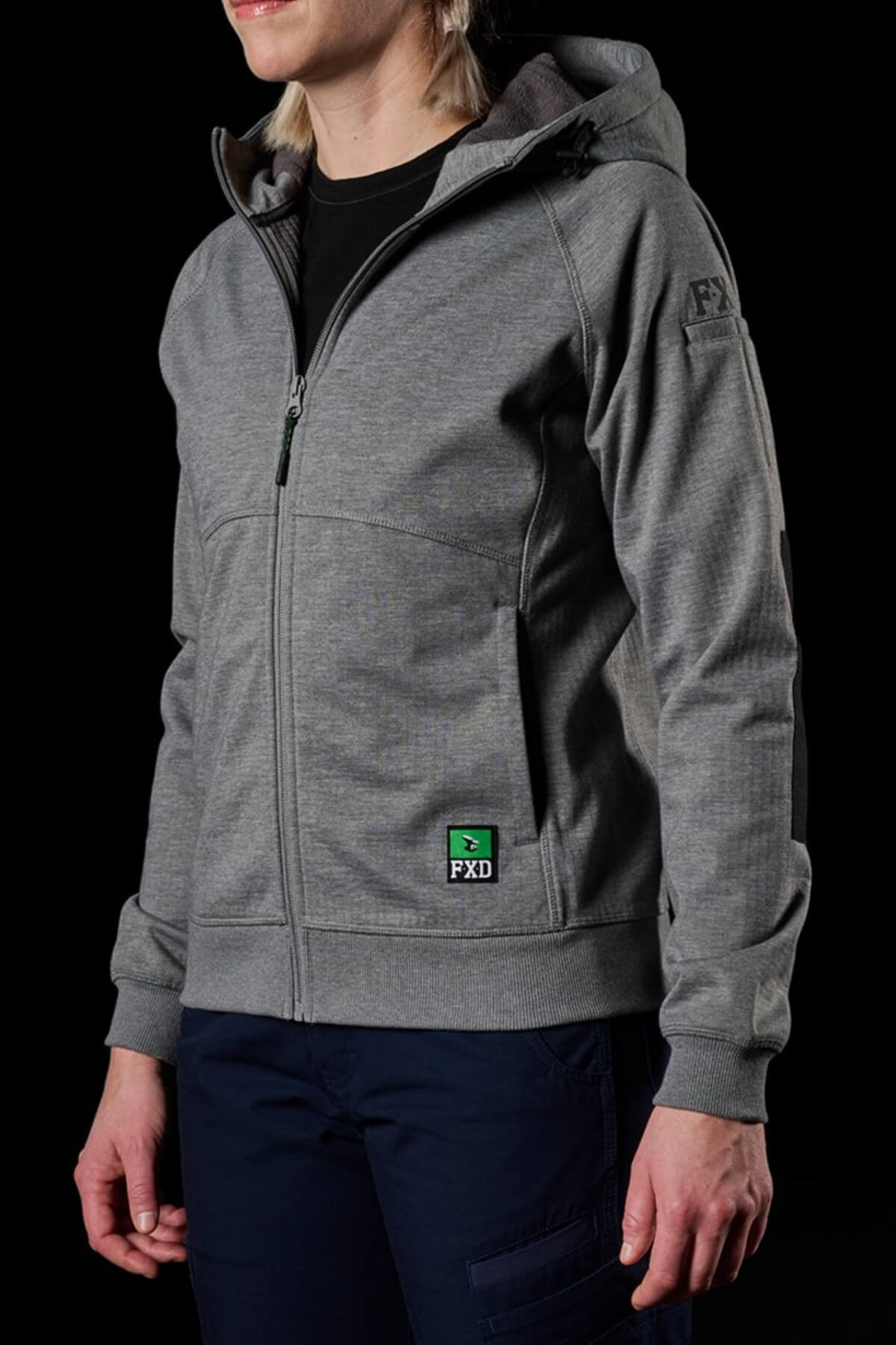 A person wearing a gray FXD Womens WF-3W Jacket with a front zipper and pockets is dressed in this windproof, water-resistant jacket that features a small green and black patch with the letters "FXD" on the lower left side. The person is also sporting dark pants, all set against a black background.